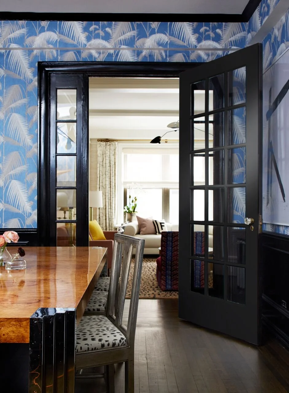 View through black french doors into a bright living room with white sofa, patterned cushions, and a window with curtains.