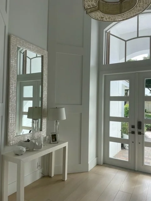 Interior of a bright entryway with a white console table, a large textured mirror, a table lamp, framed photo, and decorative objects, near glass doors leading outside.