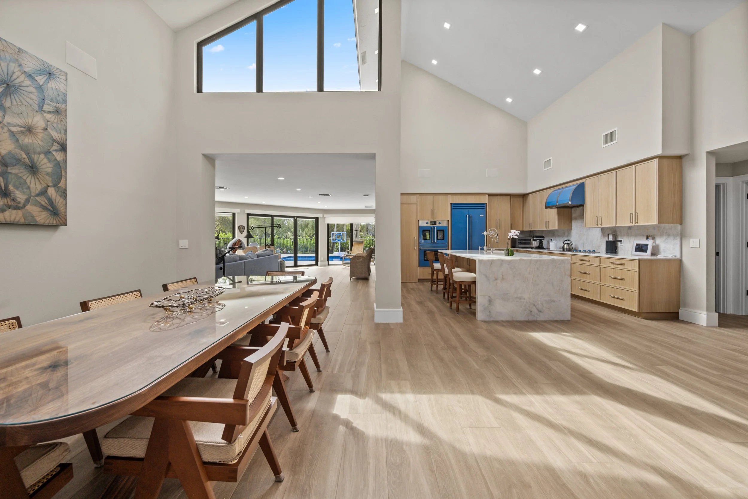 Interior view of a modern open-concept kitchen and dining area with high ceilings, large windows, wooden cabinets, a marble kitchen island, and a sliding glass door leading to an outdoor pool area.
