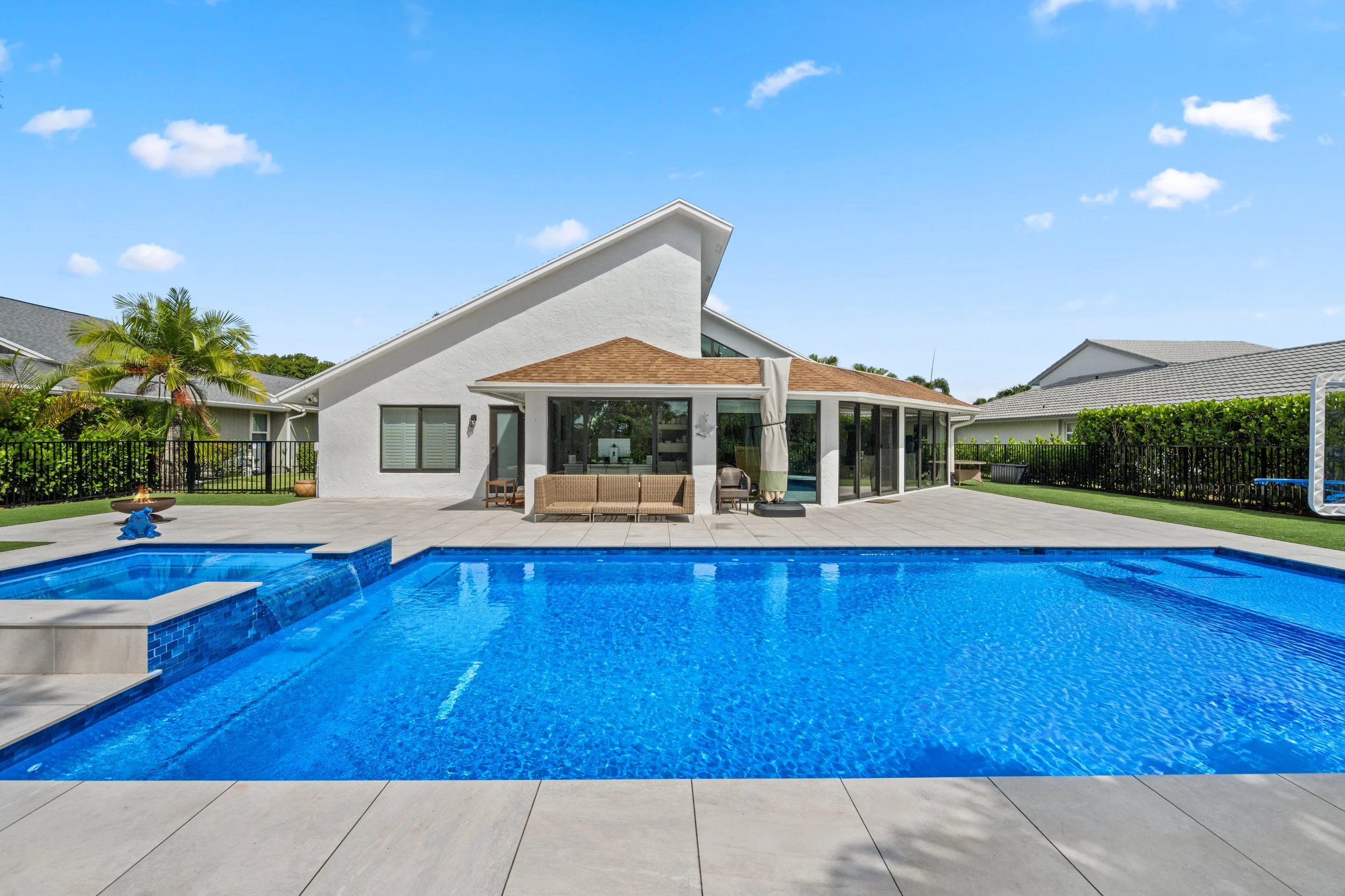 Backyard with a blue swimming pool, outdoor furniture, a white house with a unique roof design, and a clear blue sky with scattered clouds.