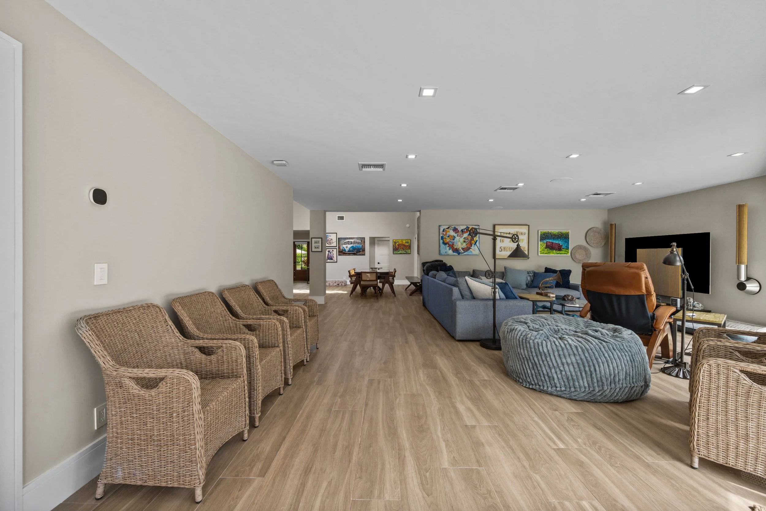 Spacious living room with beige walls, wooden flooring, multiple seating area including woven chairs and cushioned sofas, wall art, and large flat-screen TV, illuminated by recessed ceiling lights.
