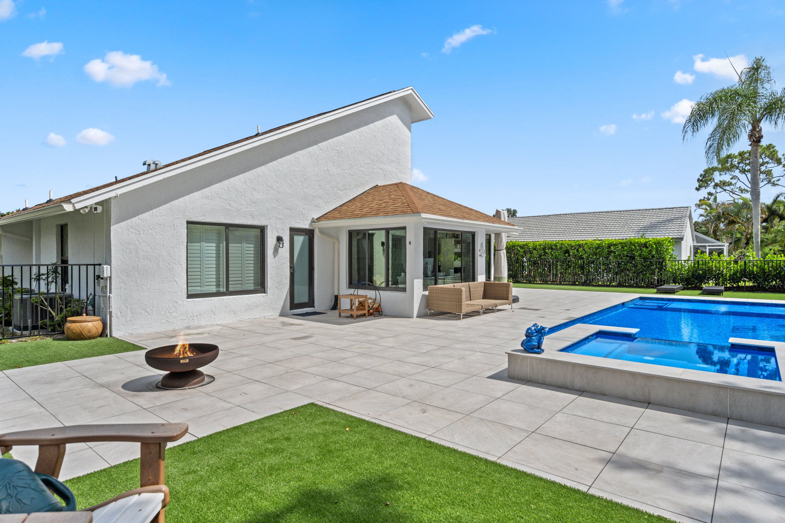 A backyard with a white stucco house, a swimming pool, outdoor seating, a firepit, and palm trees under a blue sky with clouds.