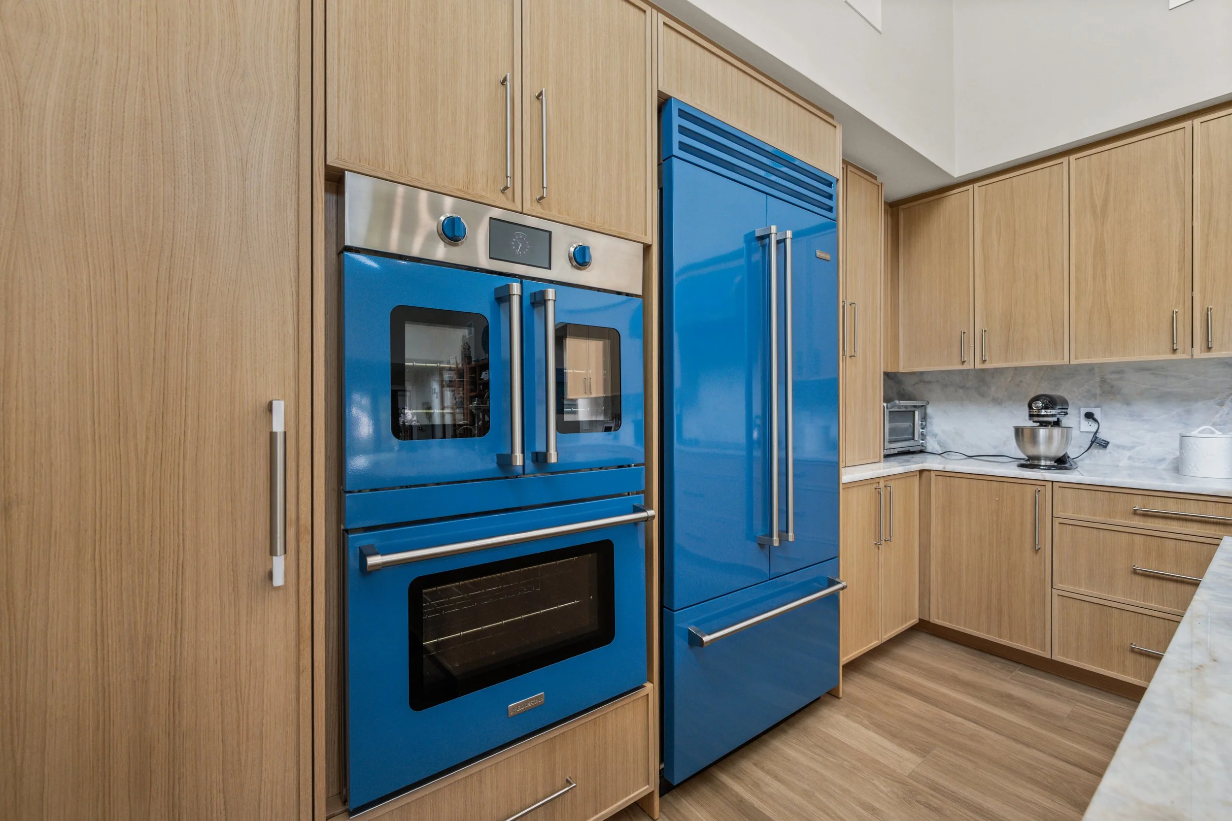 Modern kitchen with light wooden cabinets, blue double oven, blue refrigerator, marble countertop, toaster, stand mixer, and paper towel holder.
