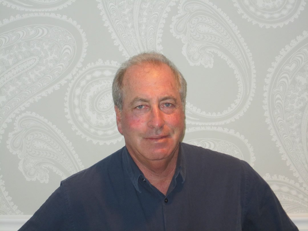 A middle-aged man with light skin, gray hair, and blue eyes, wearing a dark blue collared shirt, sitting against a light gray background with a paisley pattern.