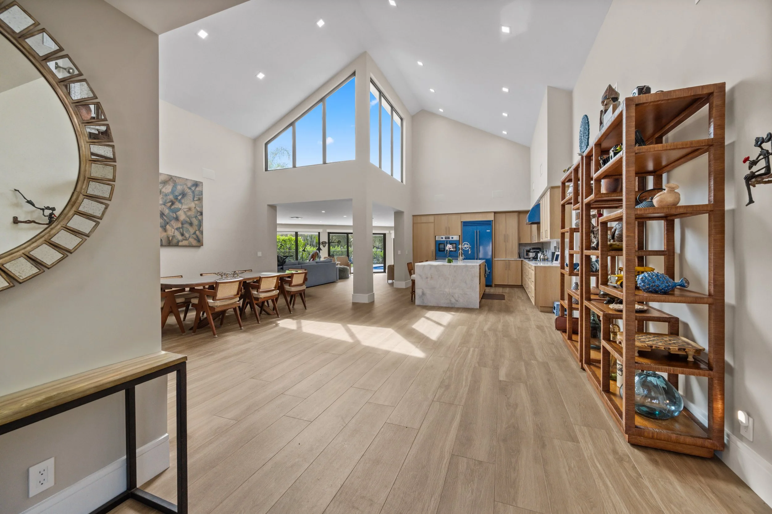 Interior view of a modern open-concept living space with high ceilings, large triangular windows, and hardwood floors. The room features a dining area with a wooden table and chairs, a kitchen with a marble island and wooden cabinetry, and a bookshel