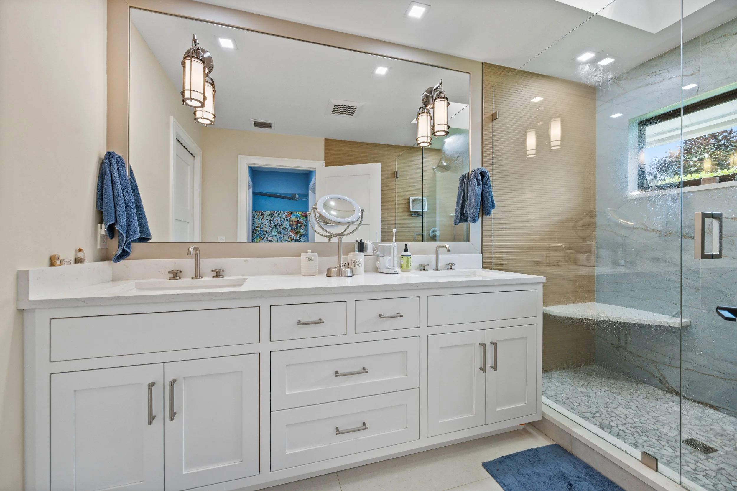 Bathroom with double sink vanity, large mirror, hanging pendant lights, blue towels, and a glass-enclosed shower with a window showing outdoor greenery.