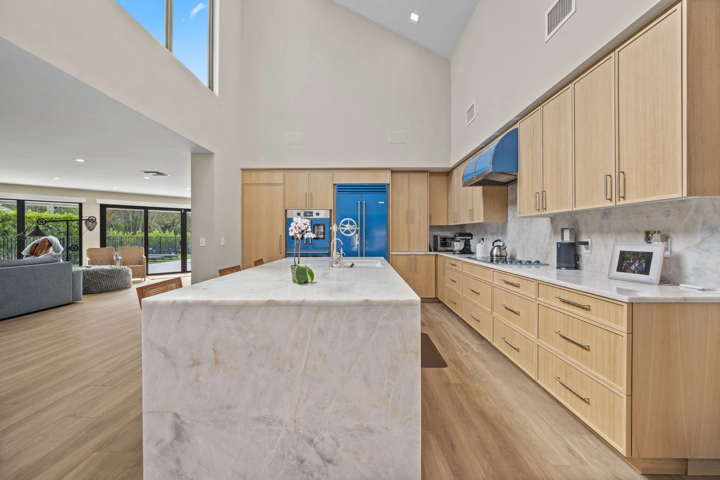 Modern kitchen with light wood cabinets, marble island, and stainless steel appliances, open to a living area with large windows and outside greenery.