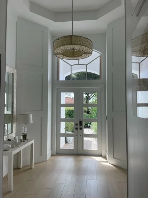 Interior view of a modern, bright entryway with a glass-paneled front door, white walls, and a hanging light fixture. There's a small table with decorative items and a window showing greenery outside.