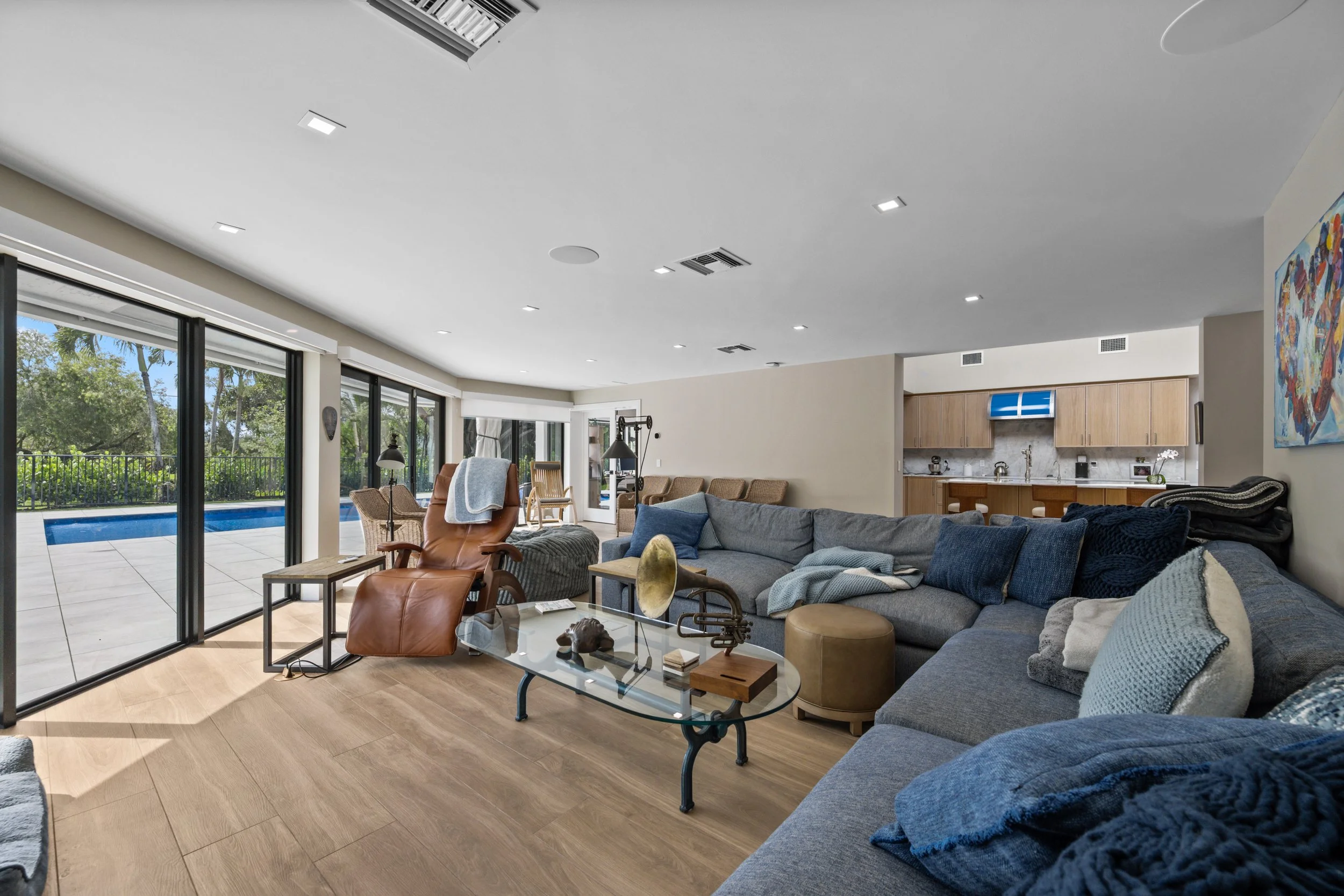 Living room with large gray sectional sofa, blue and gray throw pillows, glass coffee table, brown leather armchair, and view of backyard with pool through sliding glass doors.