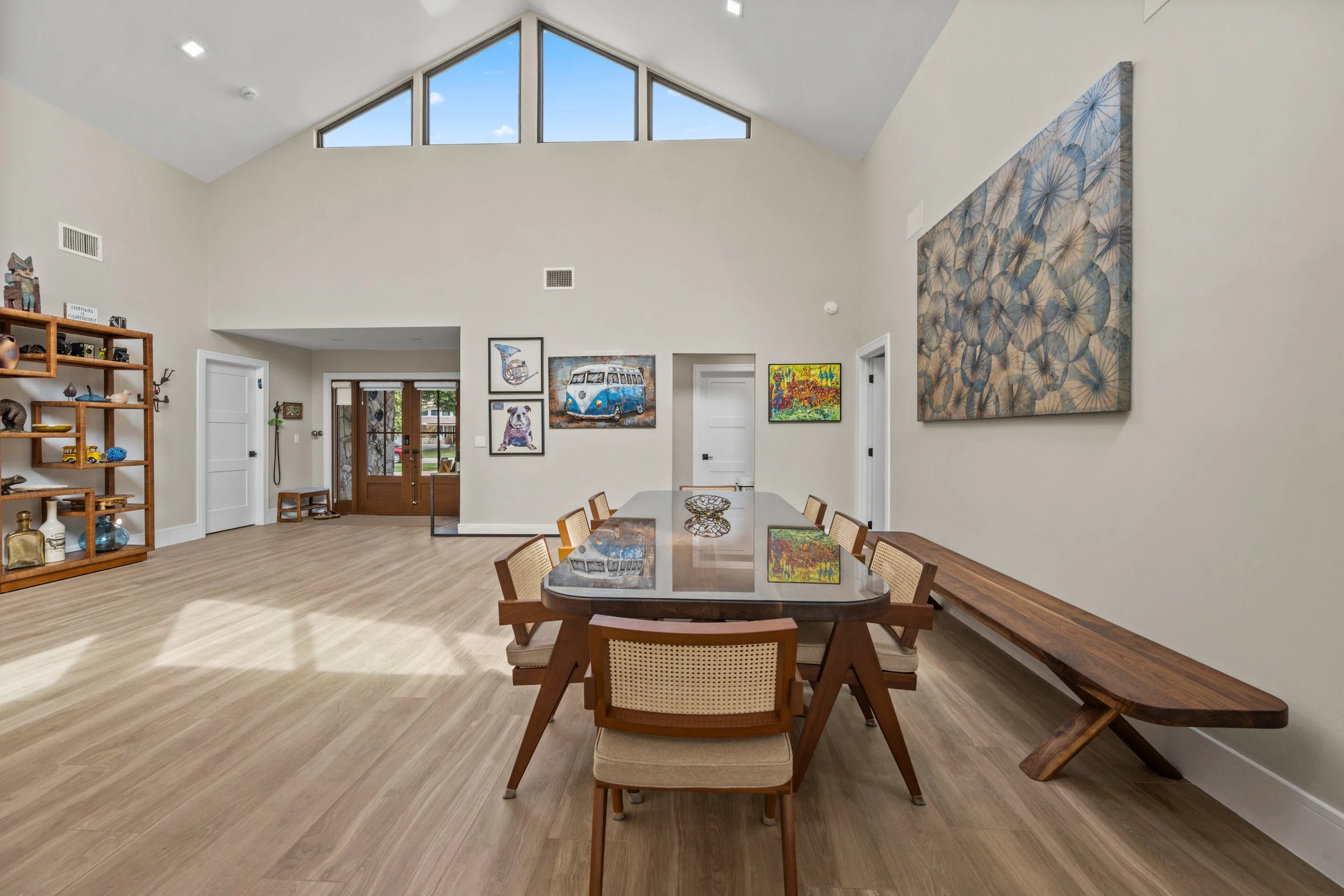 A spacious living room with high vaulted ceiling and large triangular windows at the top, allowing natural light to fill the space. The walls are decorated with colorful artwork including a painting of a Volkswagen van and other vibrant art pieces. A