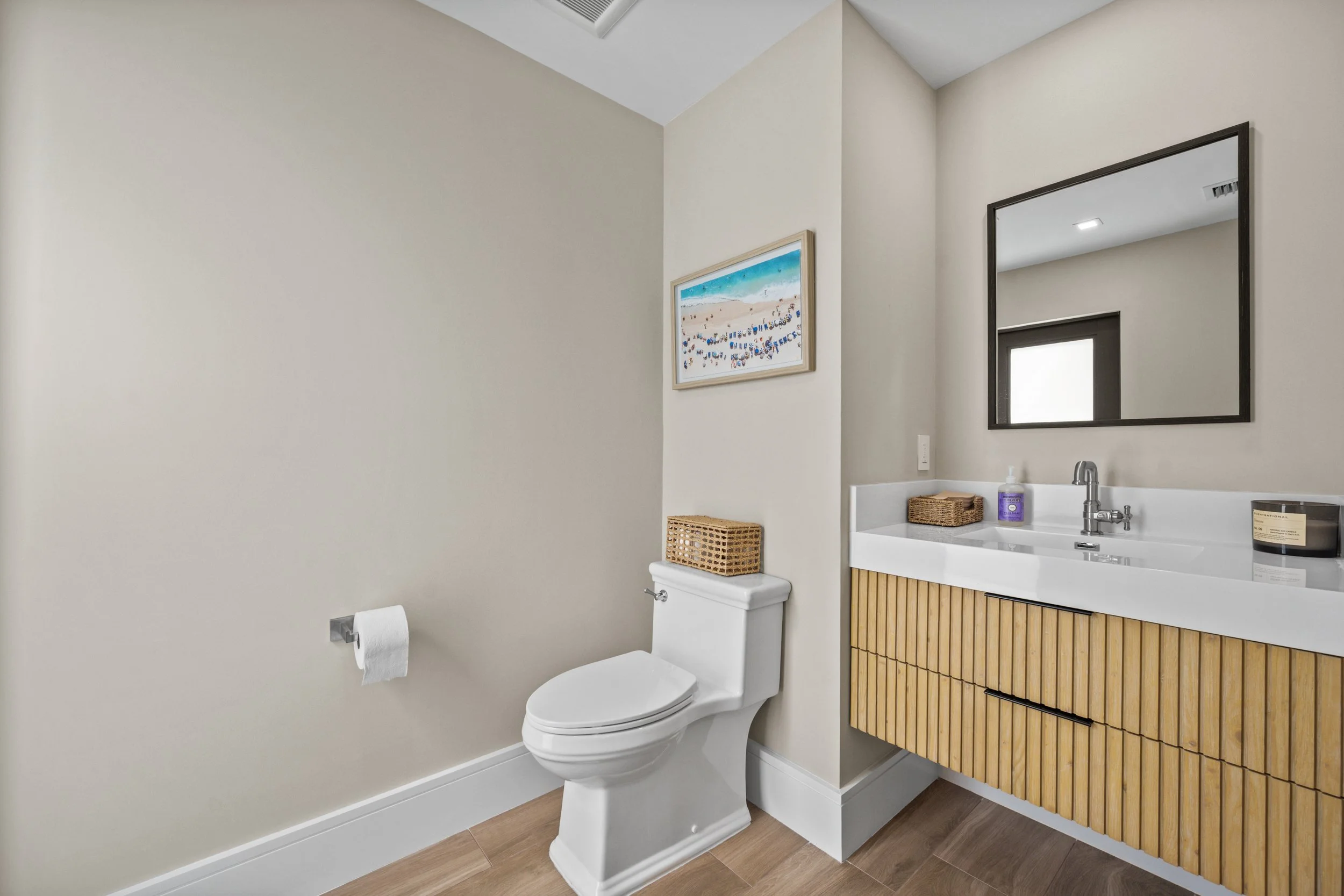 A modern bathroom featuring a white toilet with a hanging toilet paper roll, a small vanity with a wooden slatted door, a framed beach-themed painting on the wall, a sink with a silver faucet, a bottle of hand soap, and a large mirror reflecting part