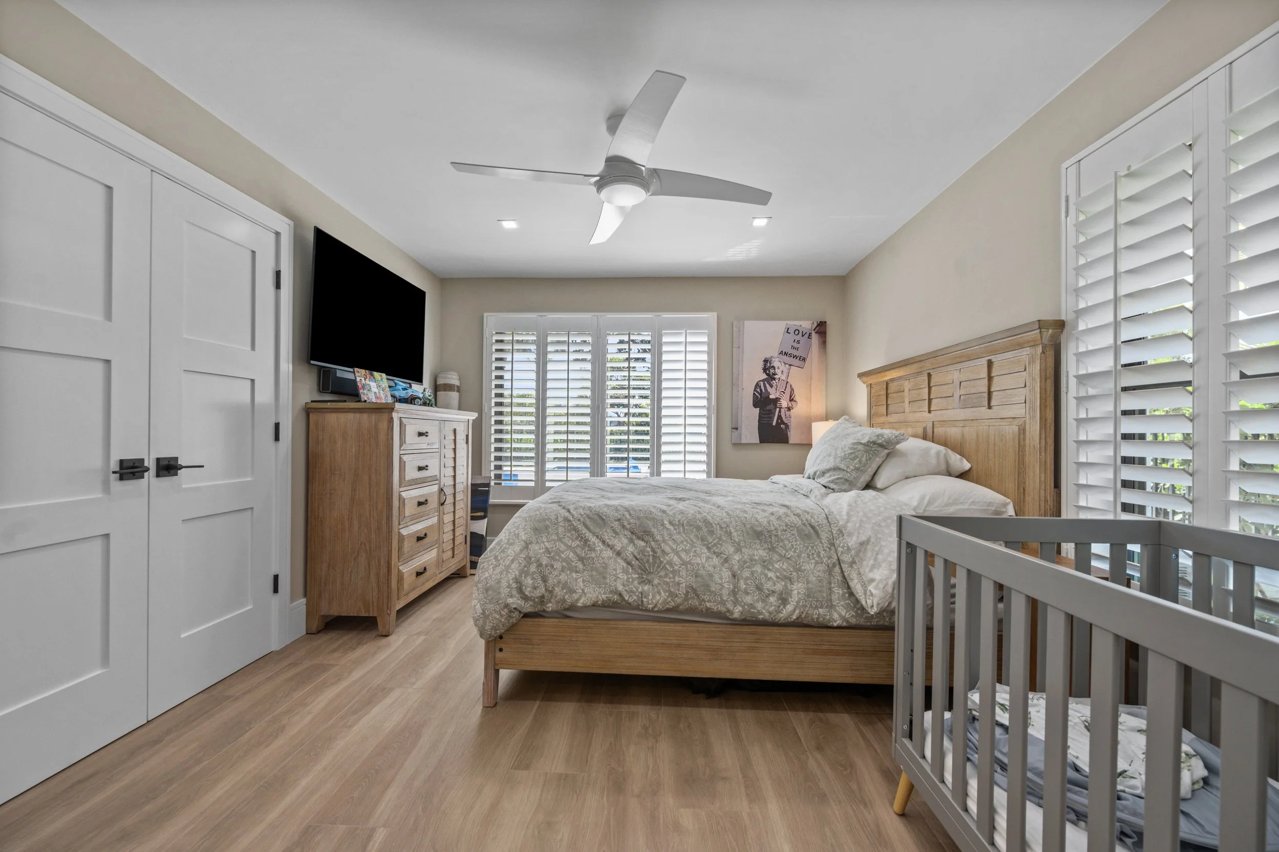 A bedroom with a wooden bed frame, white bedding, and a nursery crib. There is a wooden dresser with items on top, a wall-mounted TV, and large windows with white plantation shutters. A piece of wall art features a woman holding a sign that reads 'Lo