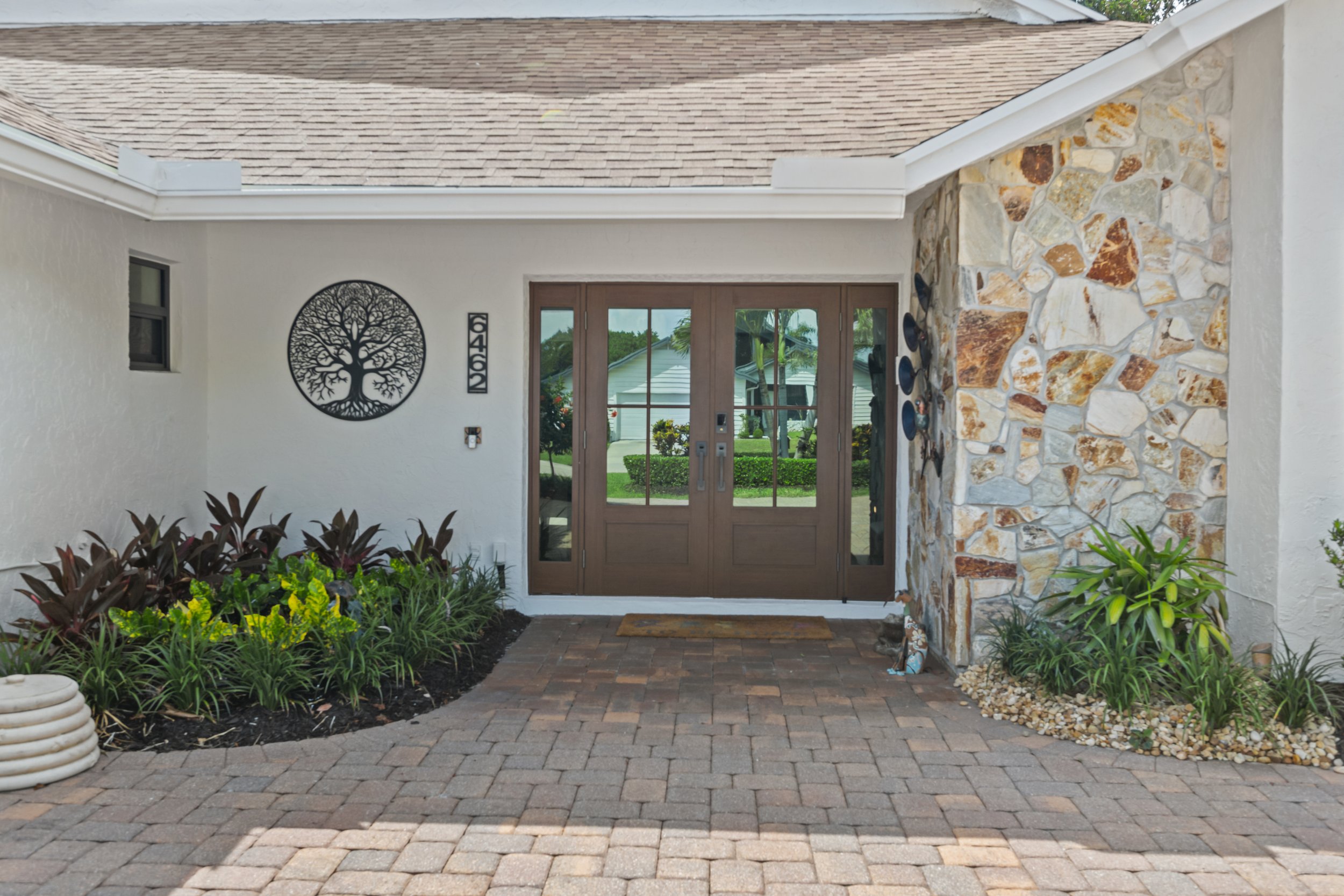 Front entrance of a house with a wooden front door, stone accents, and garden plants, potted plants, and a brick-paved walkway in the foreground.