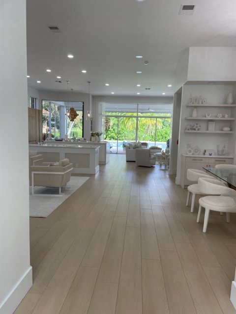 Spacious, modern living room and dining area with white furniture, built-in shelves, and large windows showing greenery outside.