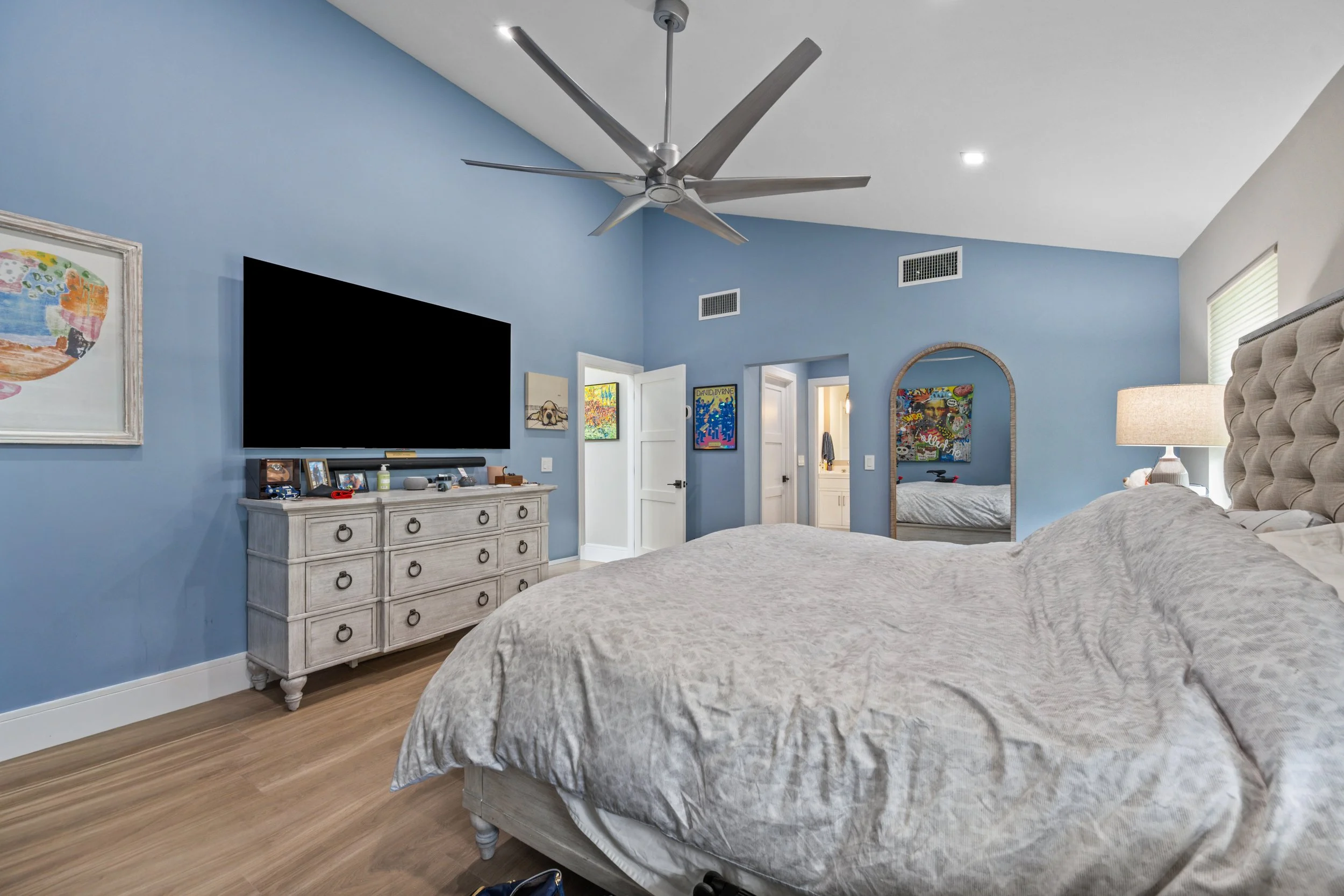 A bedroom with light blue walls, a large bed with gray and white bedding, a white dresser with framed photos, a flat-screen TV, colorful artwork on the walls, and a window with blinds.
