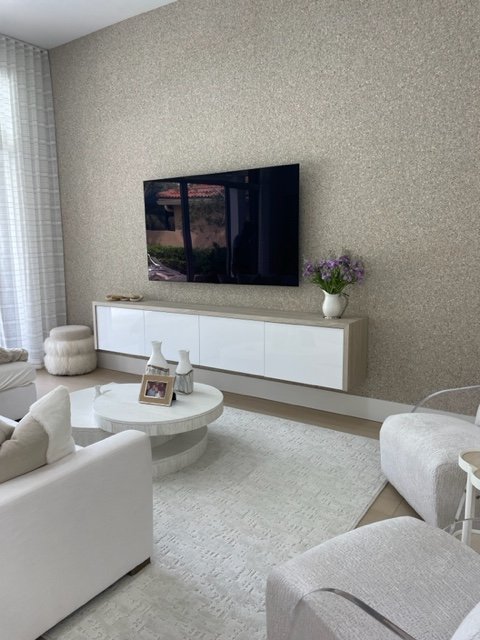 Living room with a mounted flat-screen TV on a textured beige wall, white modern entertainment console, a bouquet of purple flowers in a white vase, a white coffee table with decorative vases and picture frame, white and beige sofas, and natural ligh