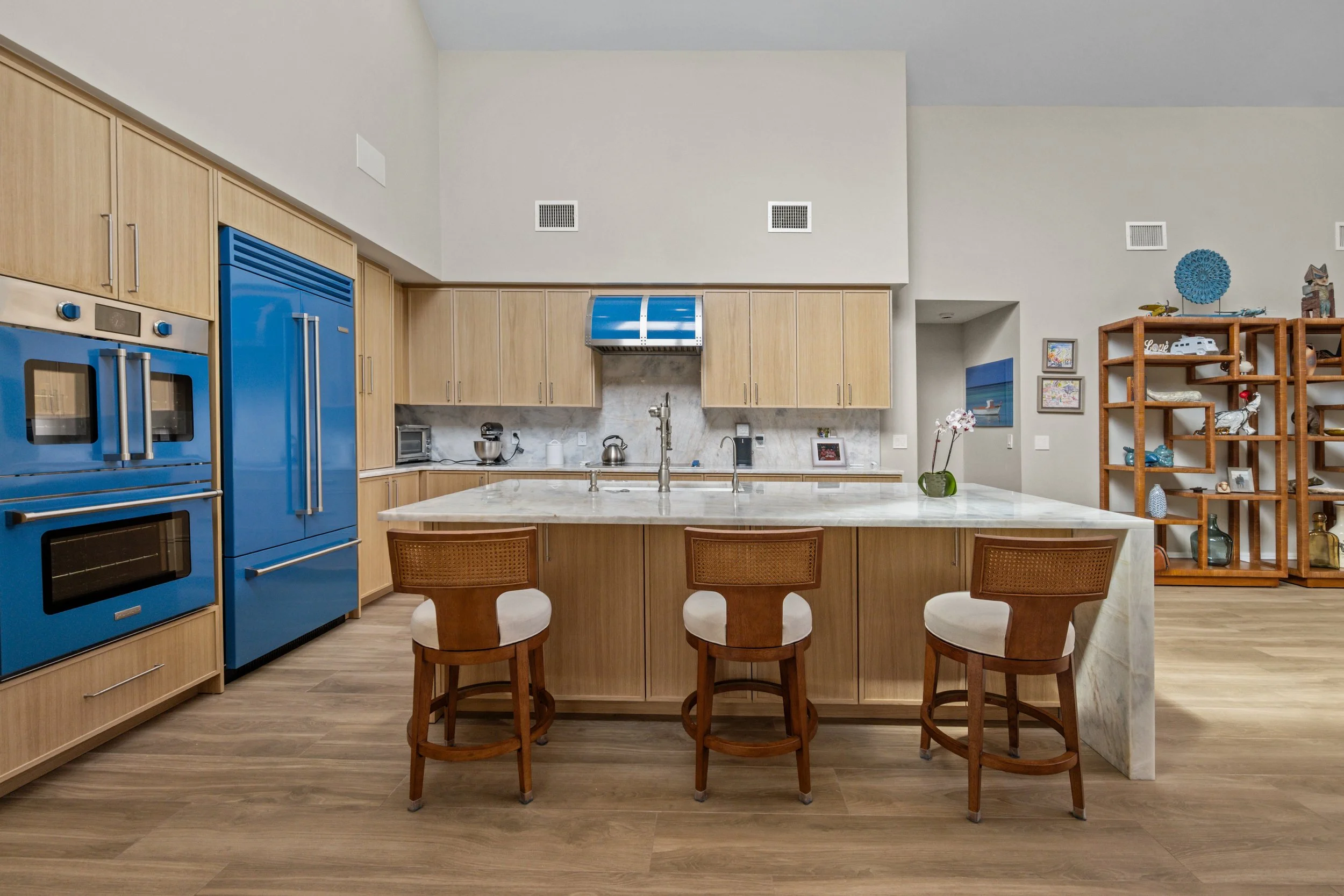 Modern kitchen with light wood cabinets, blue appliances, a marble island with three chairs, and a wooden shelving unit with decorative items and artwork.