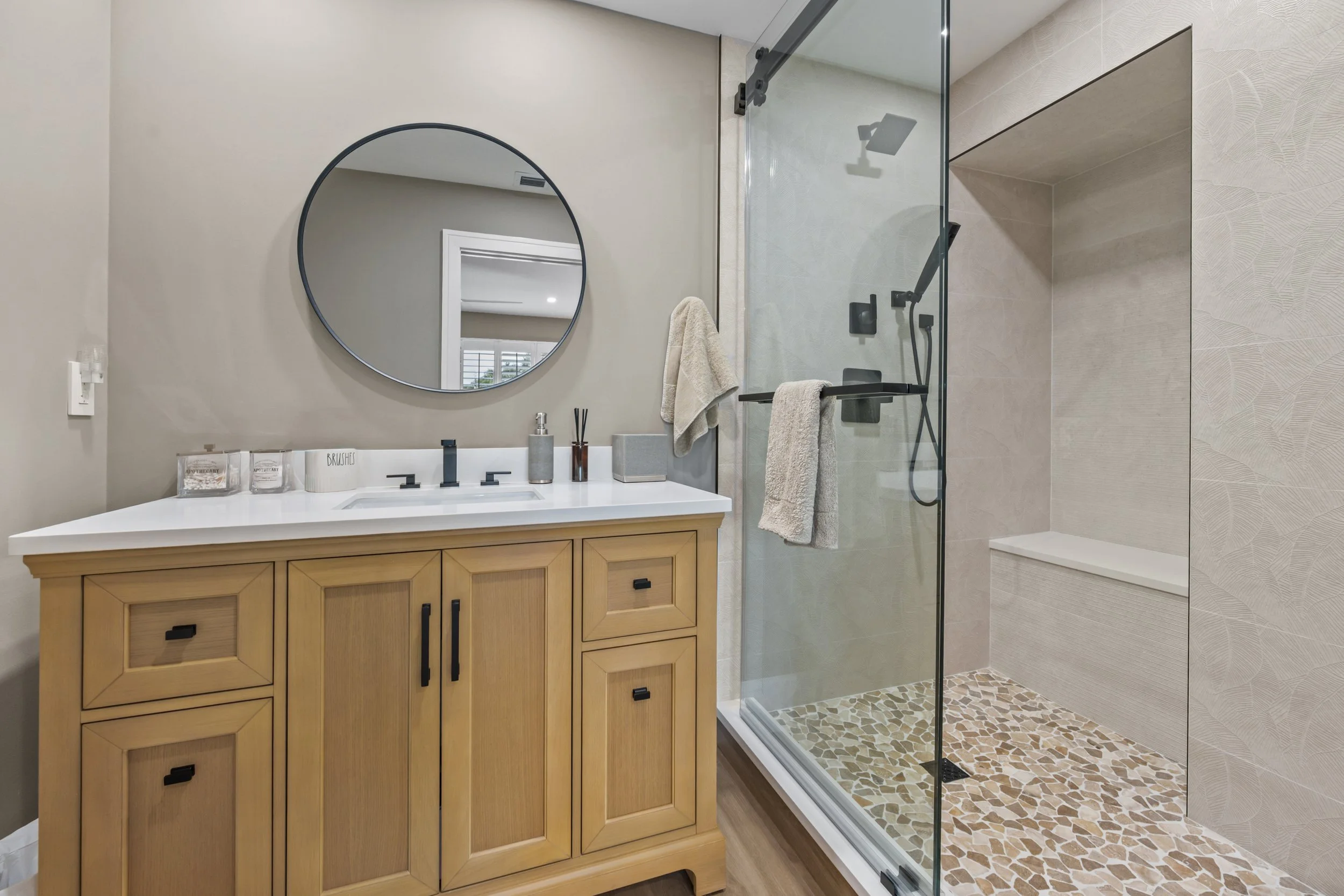 Bathroom with a wooden vanity, black hardware, a round mirror, and a glass shower with beige tile walls and pebble floor.