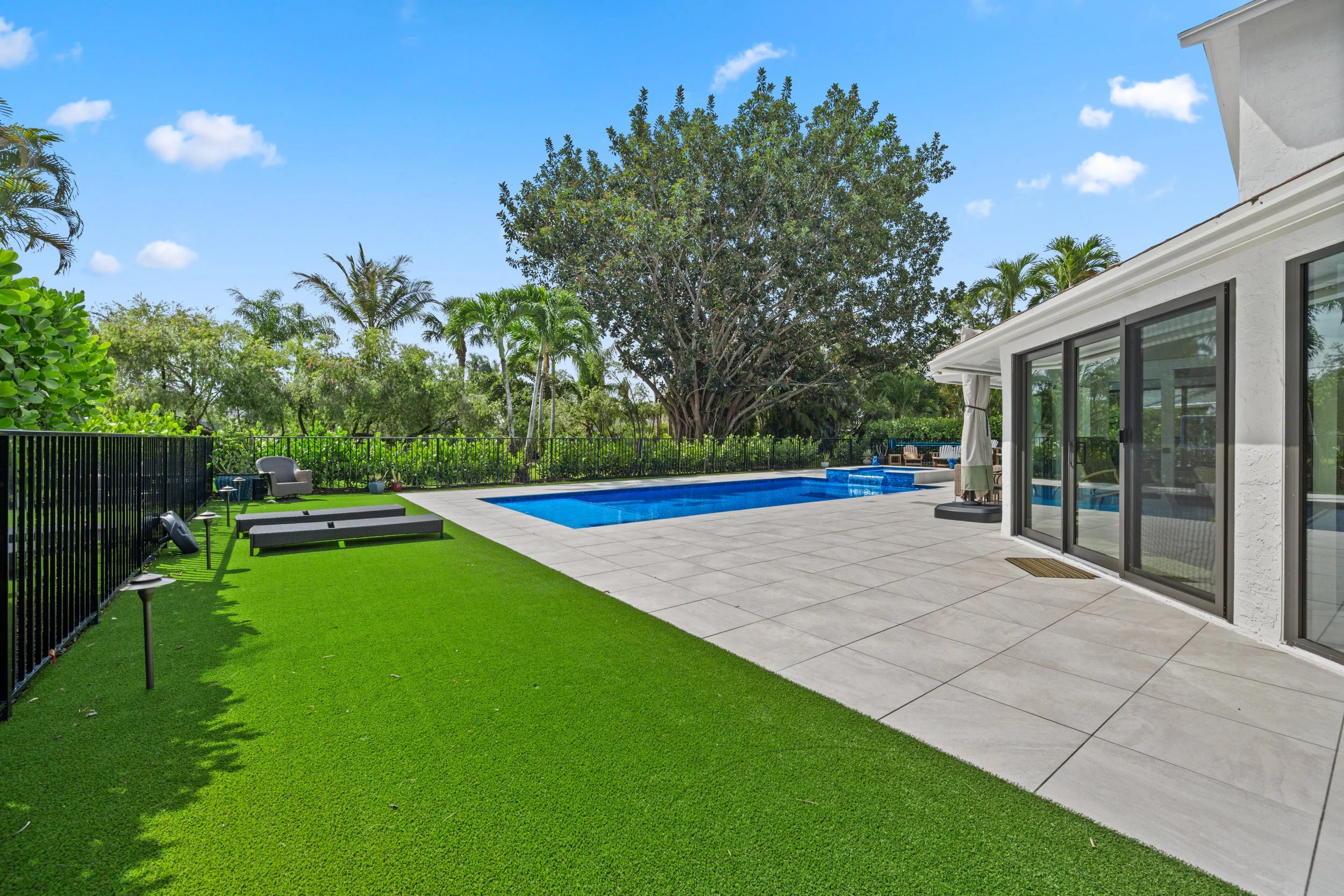 Backyard with swimming pool, green artificial grass, patio tiles, patio furniture, large tree, and house with sliding glass doors under a blue sky with clouds.