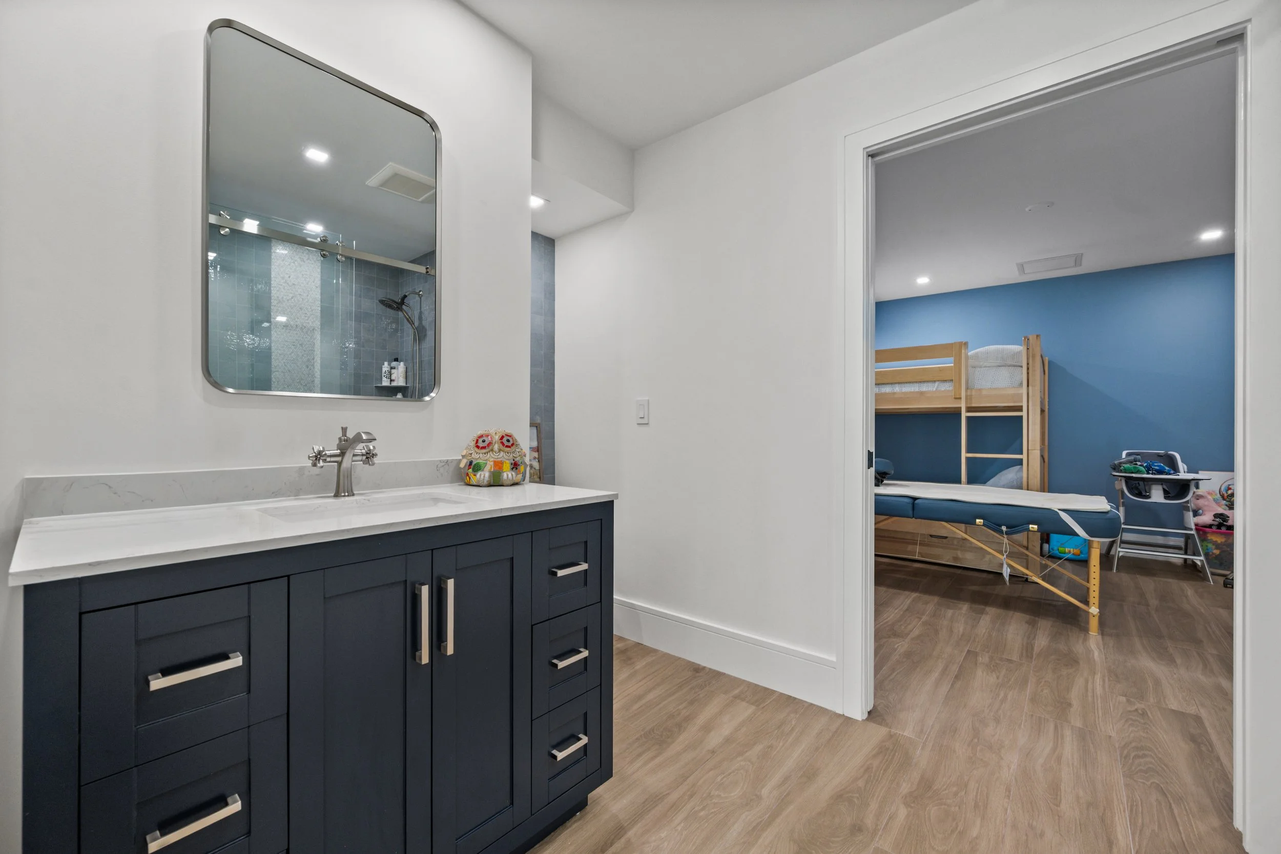 A modern bathroom with a navy blue vanity, marble countertop, and a large mirror. Beyond the doorway is a child's bedroom with a bunk bed, a blue wall, and various toys on the floor.