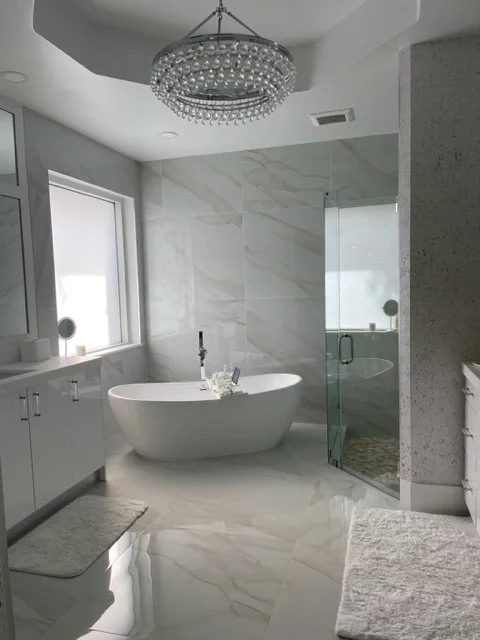 Modern bathroom with a freestanding bathtub, glass shower enclosure, white cabinets, and a large window, illuminated by a circular chandelier.