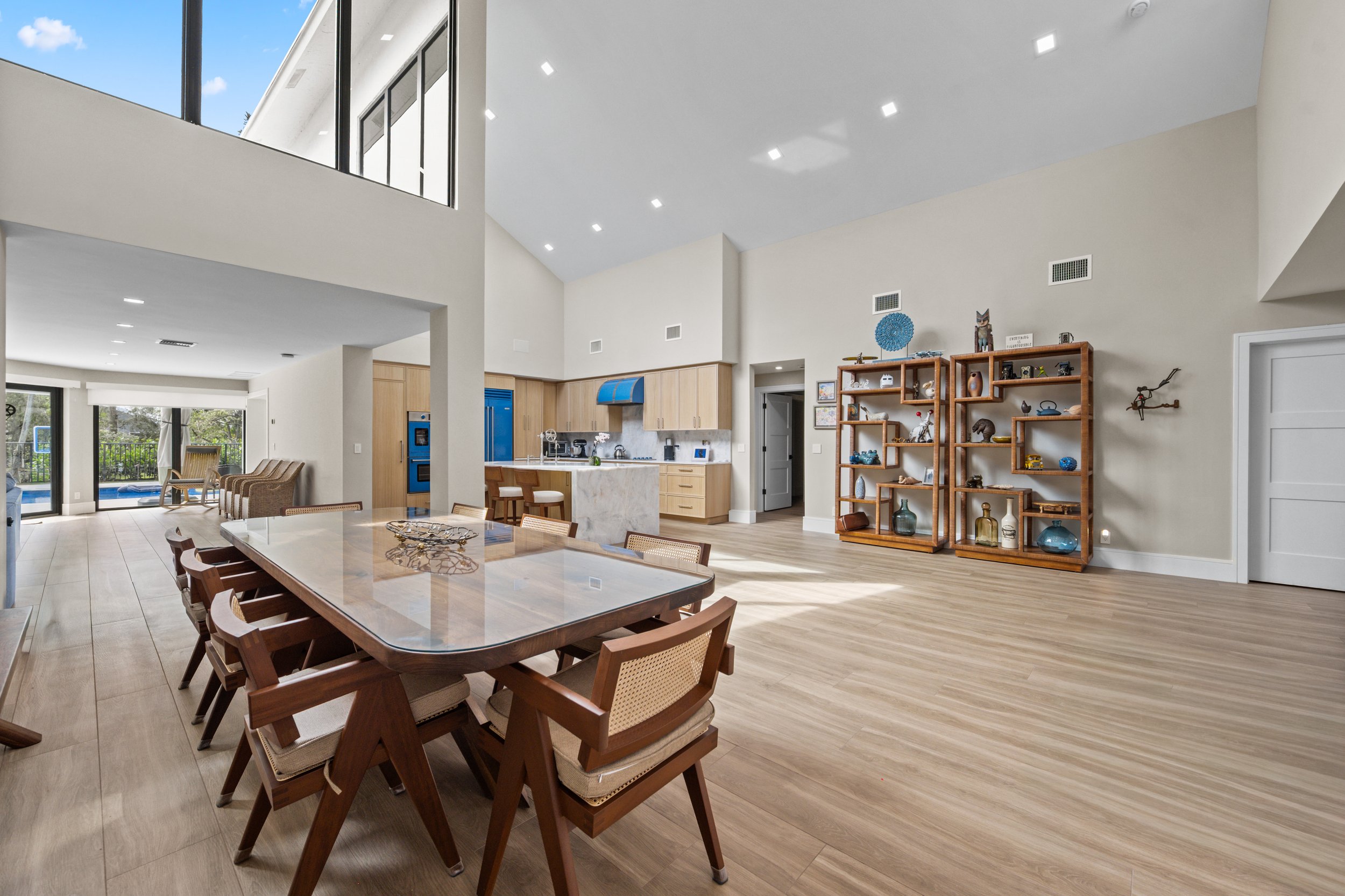 Open-concept living room and kitchen with a high ceiling, large windows, and modern decor, including a wooden shelving unit with decorative items and a large dining table with wooden chairs.