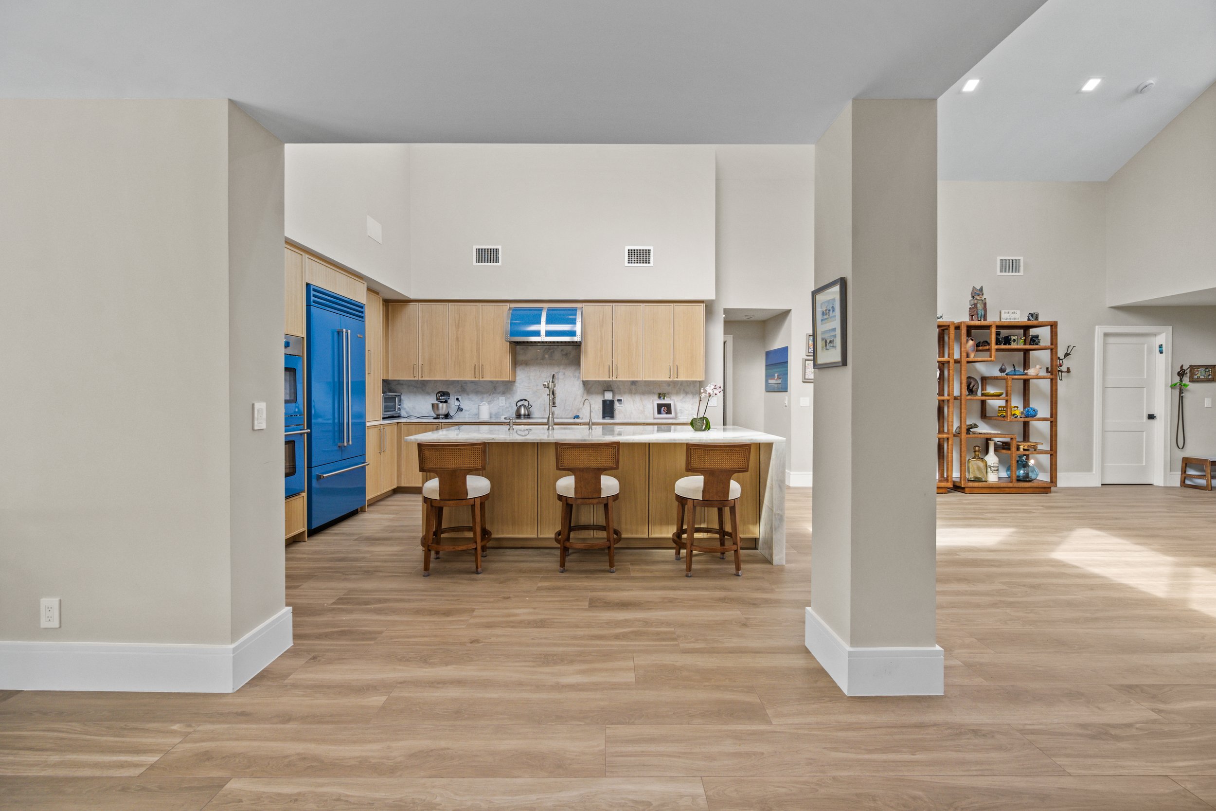 Open-concept kitchen and living space with wooden flooring, beige walls, and modern ceiling lights. The kitchen has light wood cabinets, a blue refrigerator, and a marble countertop island with three wooden stools. The living area features a wooden s