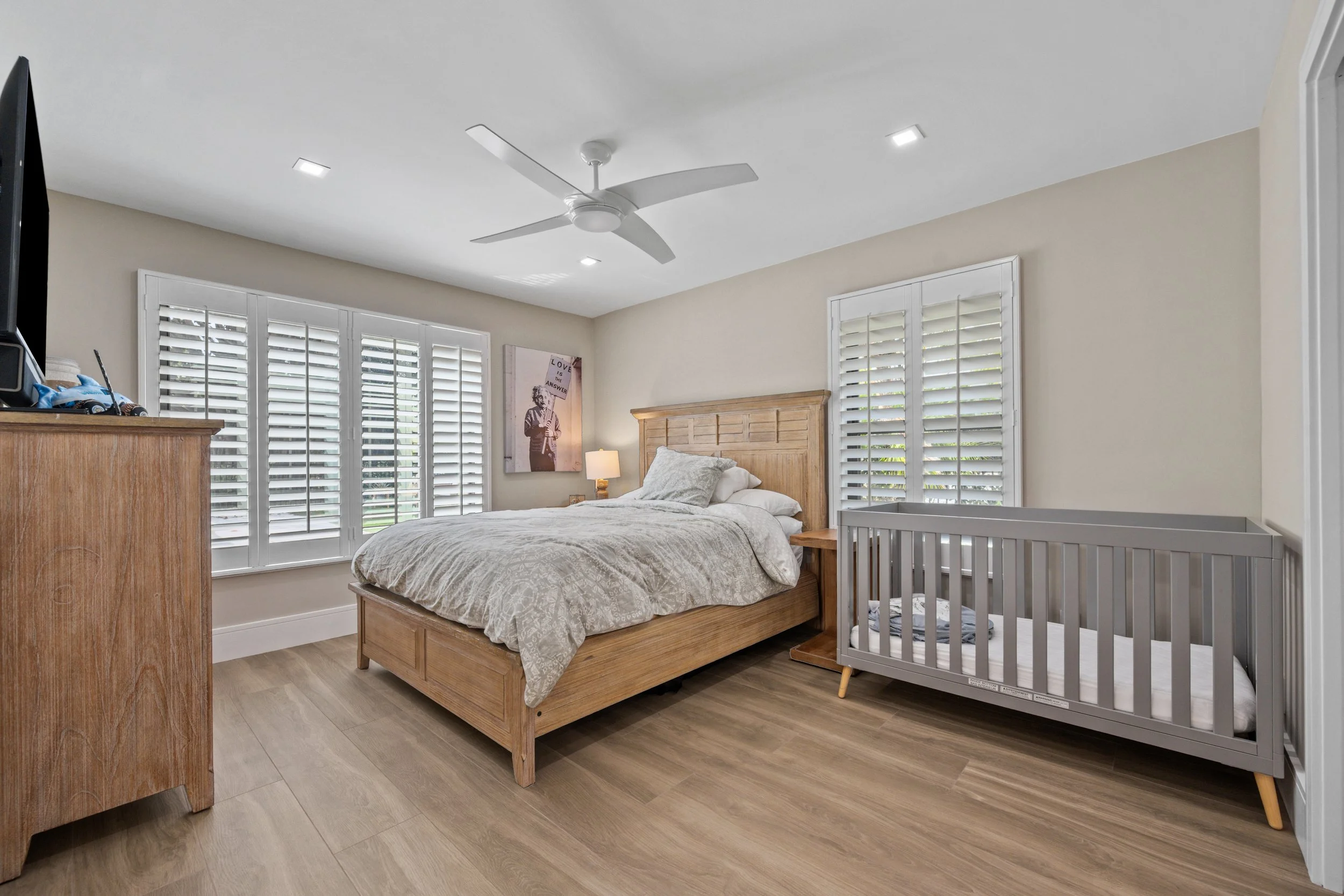 A bedroom with a wooden bed frame, beige bedding, and windows with white plantation shutters. There is a ceiling fan, a nightstand with a lamp, a piece of wall art, and a baby crib.