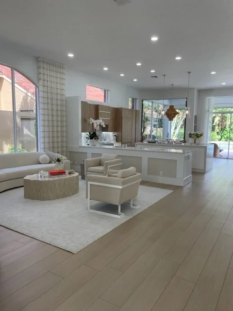 Bright, modern open-concept living room and kitchen with white and beige decor, large windows, and hardwood floors.