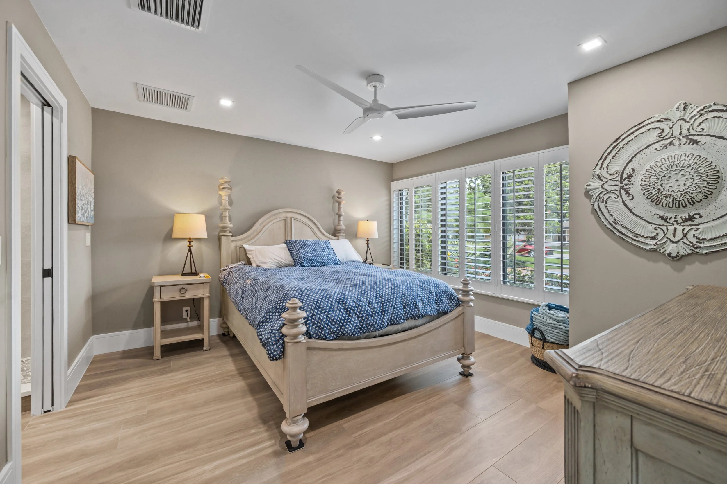 A cozy bedroom with a white wooden bed frame, blue and white bedding, two nightstands with lamps, large windows with white shutters, and beige walls. A ceiling fan hangs from the ceiling.