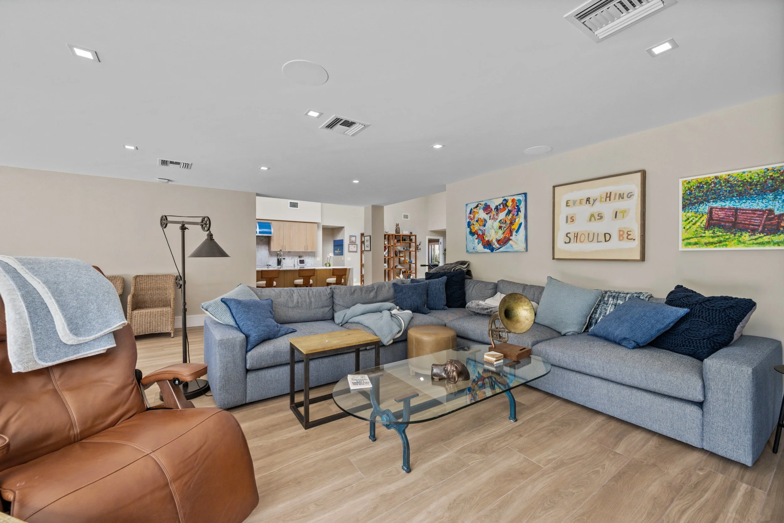 Living room with a large gray sectional sofa with blue and gray pillows, a glass coffee table, and colorful artwork on the walls. There is a brown leather armchair in the foreground and a wicker chair in the background. The room has wood flooring and