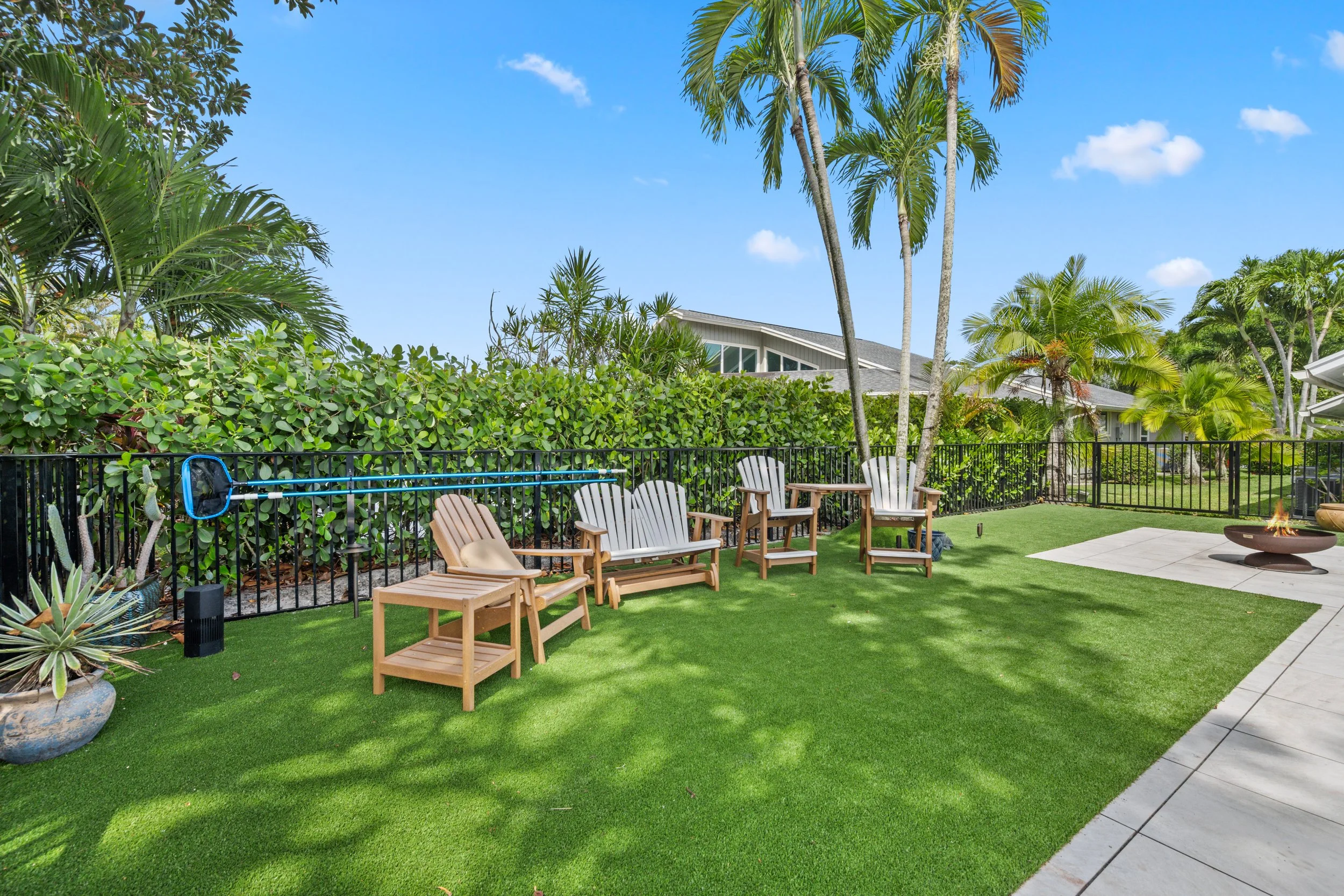 Backyard patio with wooden chairs, a firepit, lush green bushes, tall palm trees, on a sunny day with a blue sky.