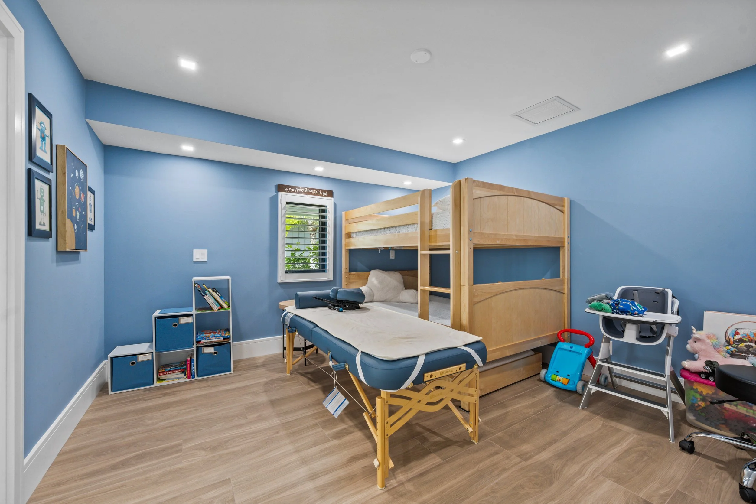 A children's bedroom with blue walls, a window with horizontal blinds, and a wall decorated with framed space theme artwork. It features a natural wood bunk bed, a foldable cot, and various toys and books around the room.