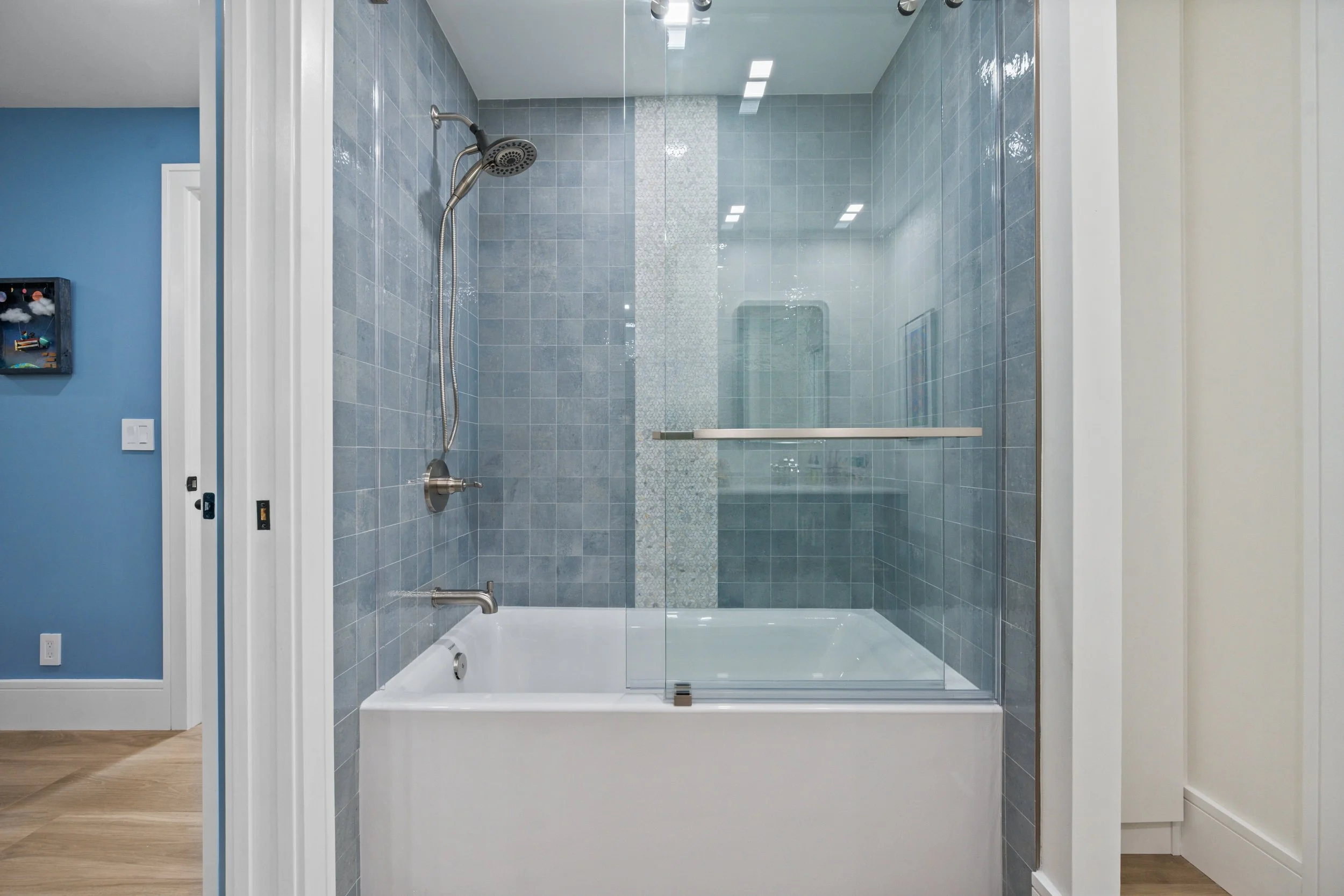 Glass shower enclosure with blue tiled walls, a hand-held shower head, and a chrome handle, situated in a modern bathroom with blue and white walls and wood flooring.