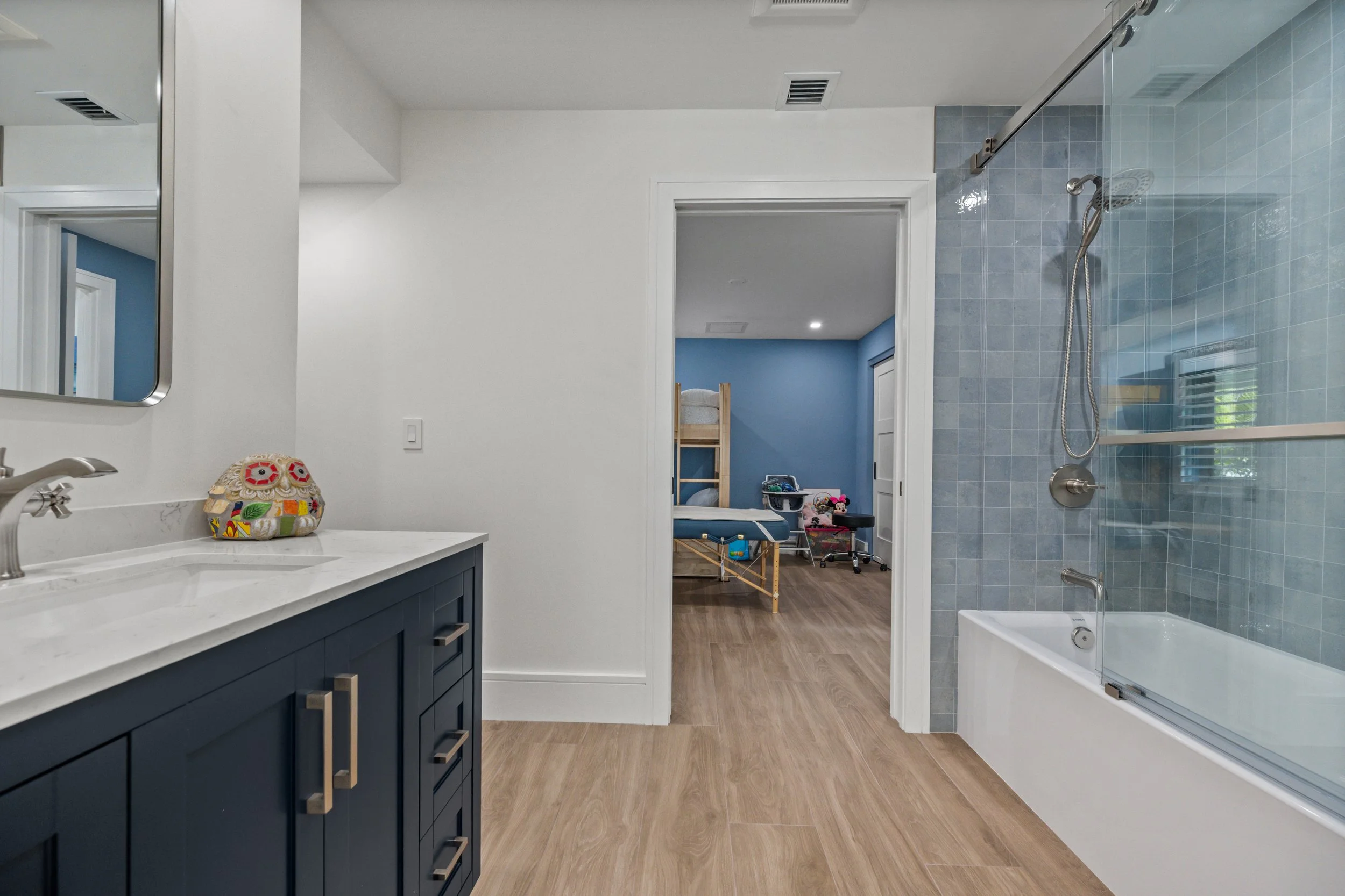 Modern bathroom with navy blue cabinetry, white marble countertop, and a large mirror. The room has wood flooring, a walk-in shower with blue tiles, and a view into an adjacent kids' bedroom with a blue wall, children’s bed, toys, and a changing tabl