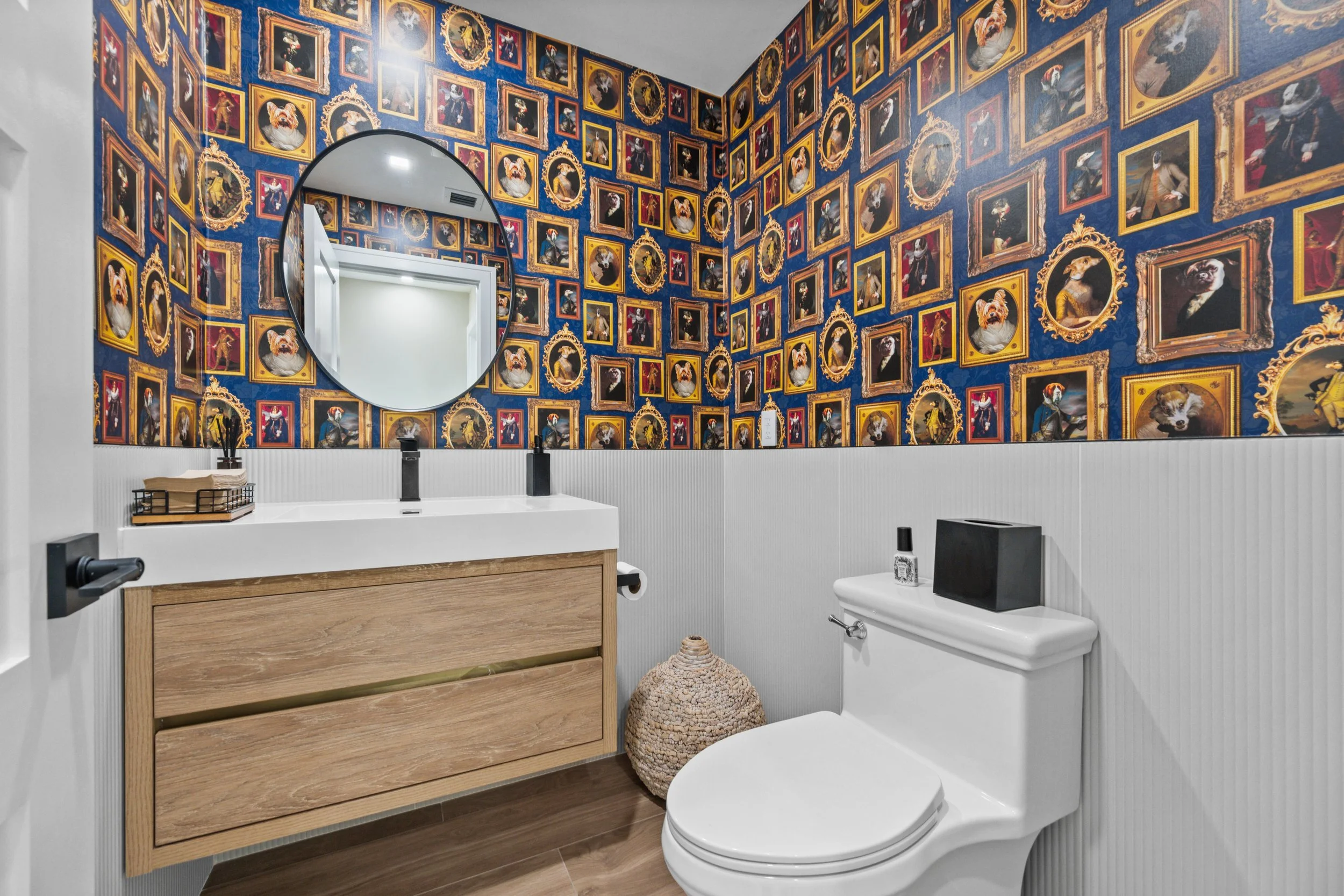 Bathroom with blue wallpaper covered in framed photos of dogs dressed as royalty, large round mirror, white sink vanity with black faucet, toilet with black paper holder, woven basket, and small black speaker on the toilet tank.