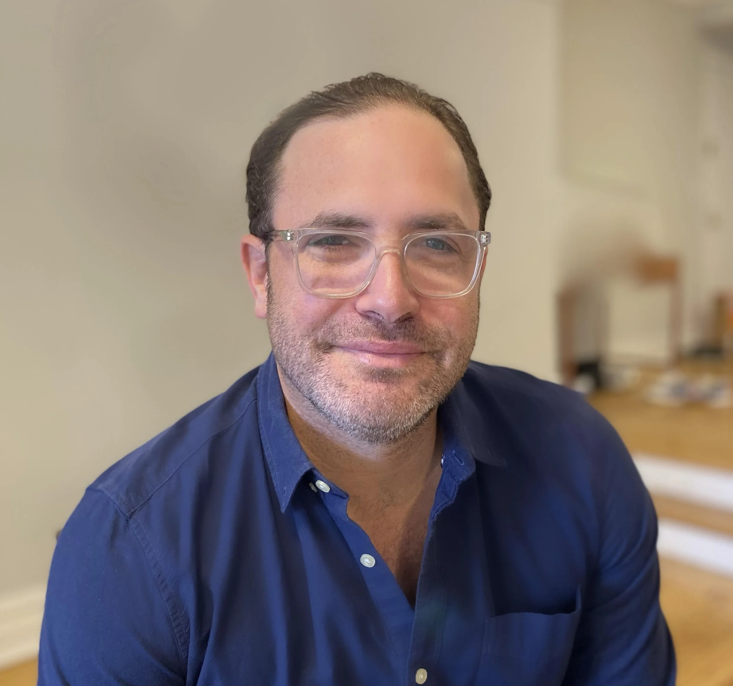 Close-up of a smiling man with glasses wearing a blue shirt, indoors with a neutral background.