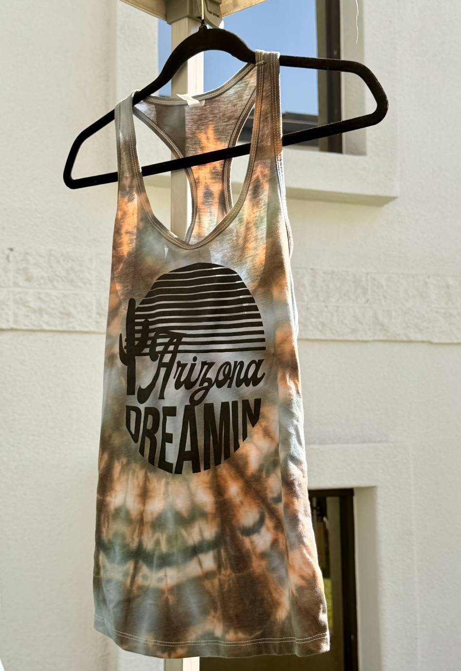 Tie-dye tank top with a cactus and sunset graphic and the words 'Arizona Dreamin' hanging on a black hanger in front of a white wall.
