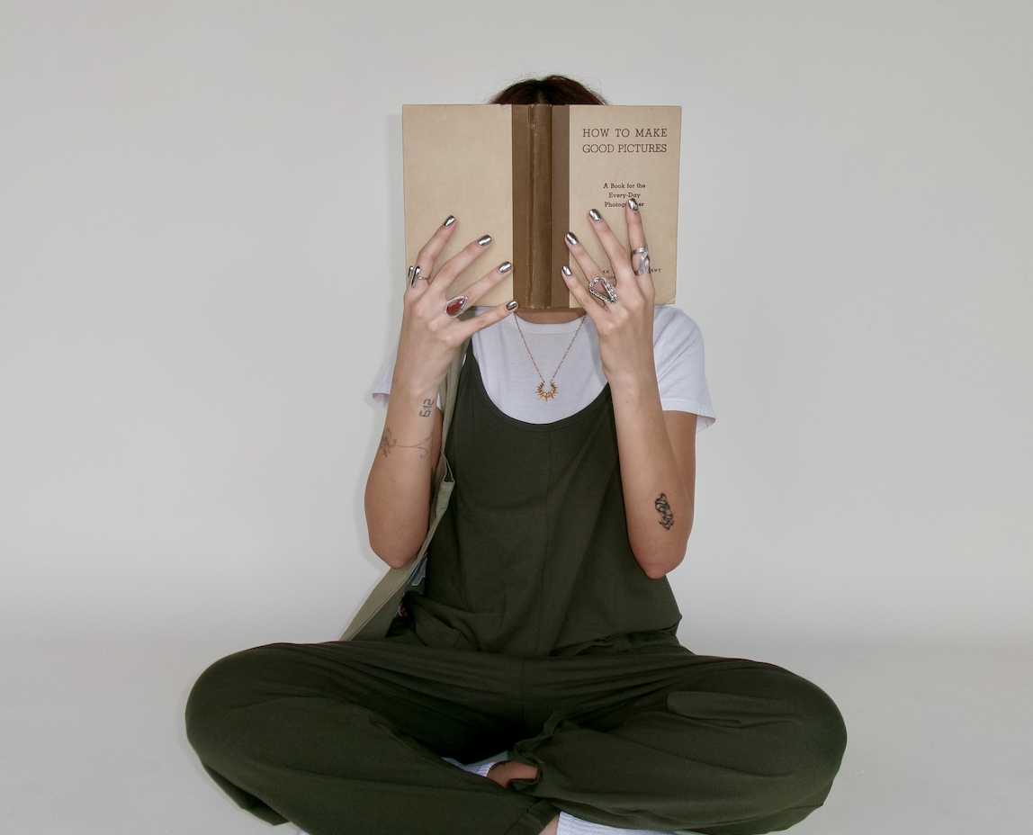Person sitting cross-legged against a plain white wall, reading a book titled 'How to Make Good Pictures', with their face hidden behind the book