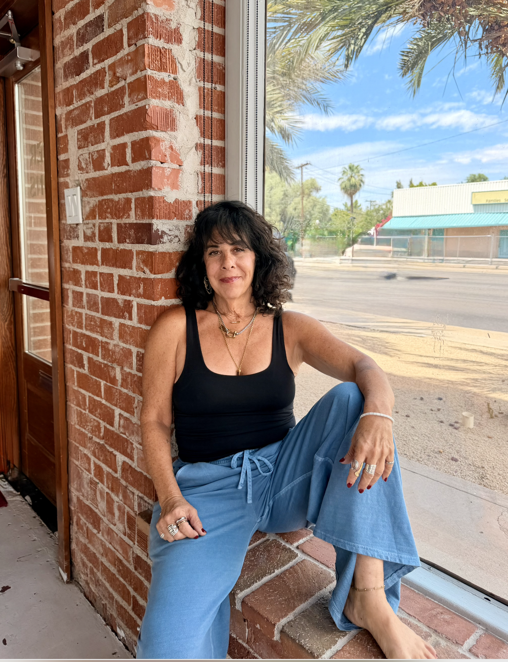 Woman sitting cross-legged on a brick window ledge inside a building with a brick wall, wearing a black tank top and blue wide-leg pants, jewelry, and with a window showing palm trees and a blue sky outside.