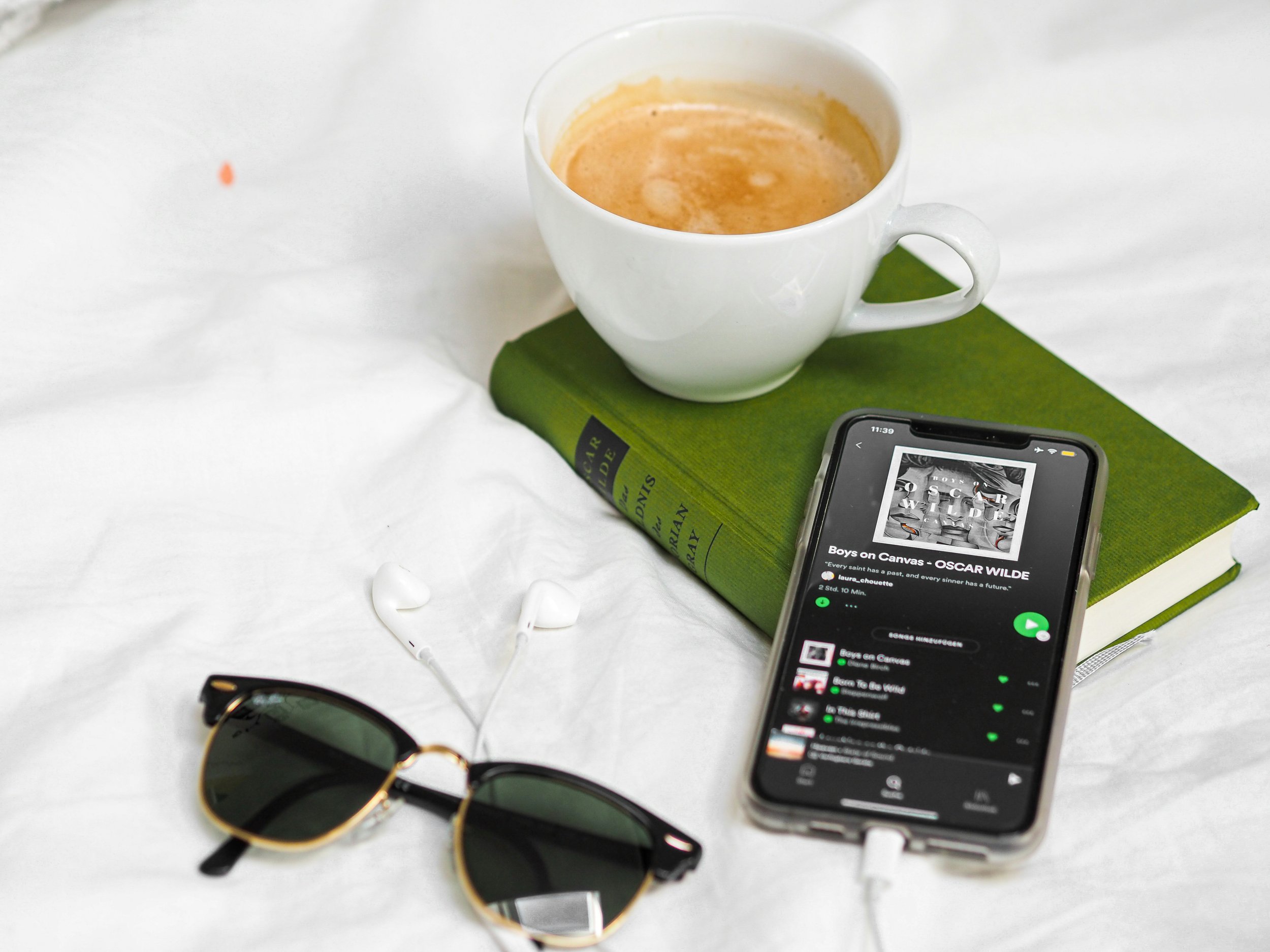 A white coffee cup filled with a light brown beverage resting on a green book, with a pair of black sunglasses, a smartphone displaying a music playlist, and some white earphones on a white surface.