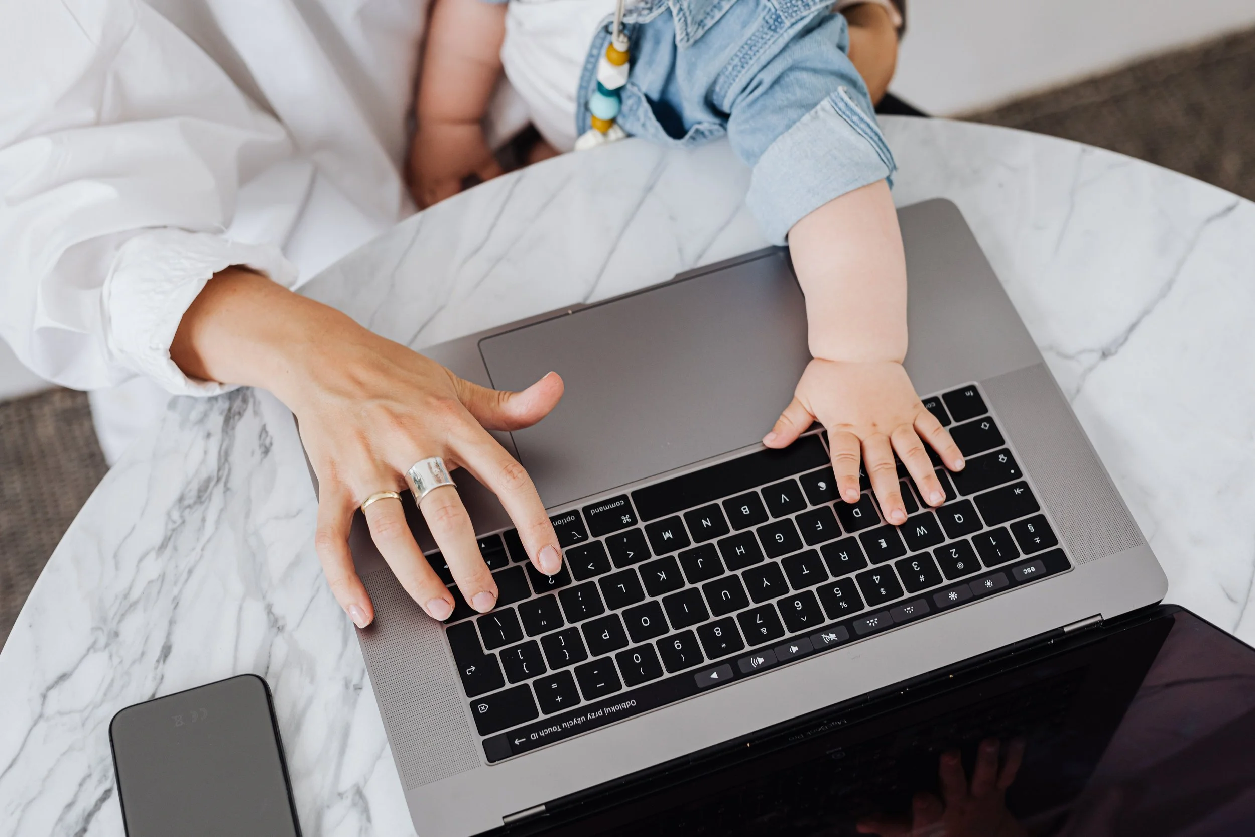 A child's hand on a laptop keyboard with an adult's hand guiding. The child is wearing a denim jacket, and the setting is a marble table.
