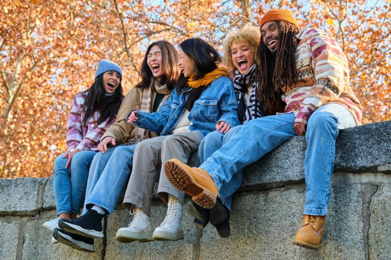 Group of five friends sitting on a stone wall, laughing and enjoying each other's company amid autumn foliage.