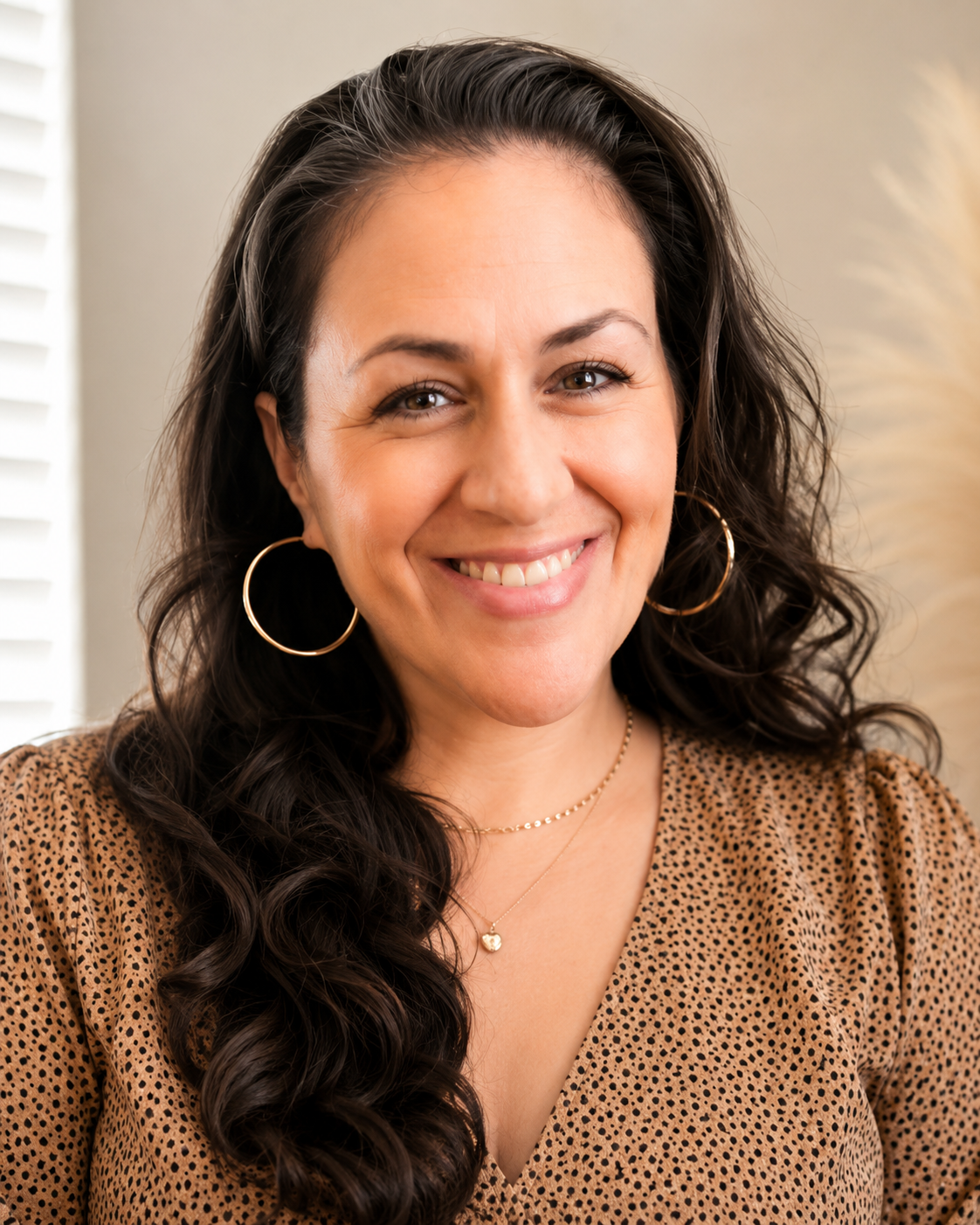 A woman with long, dark, curly hair smiling at the camera. She is wearing gold hoop earrings, a delicate gold necklace with a heart pendant, and a brown blouse with black polka dots. There are white blinds on the window behind her.