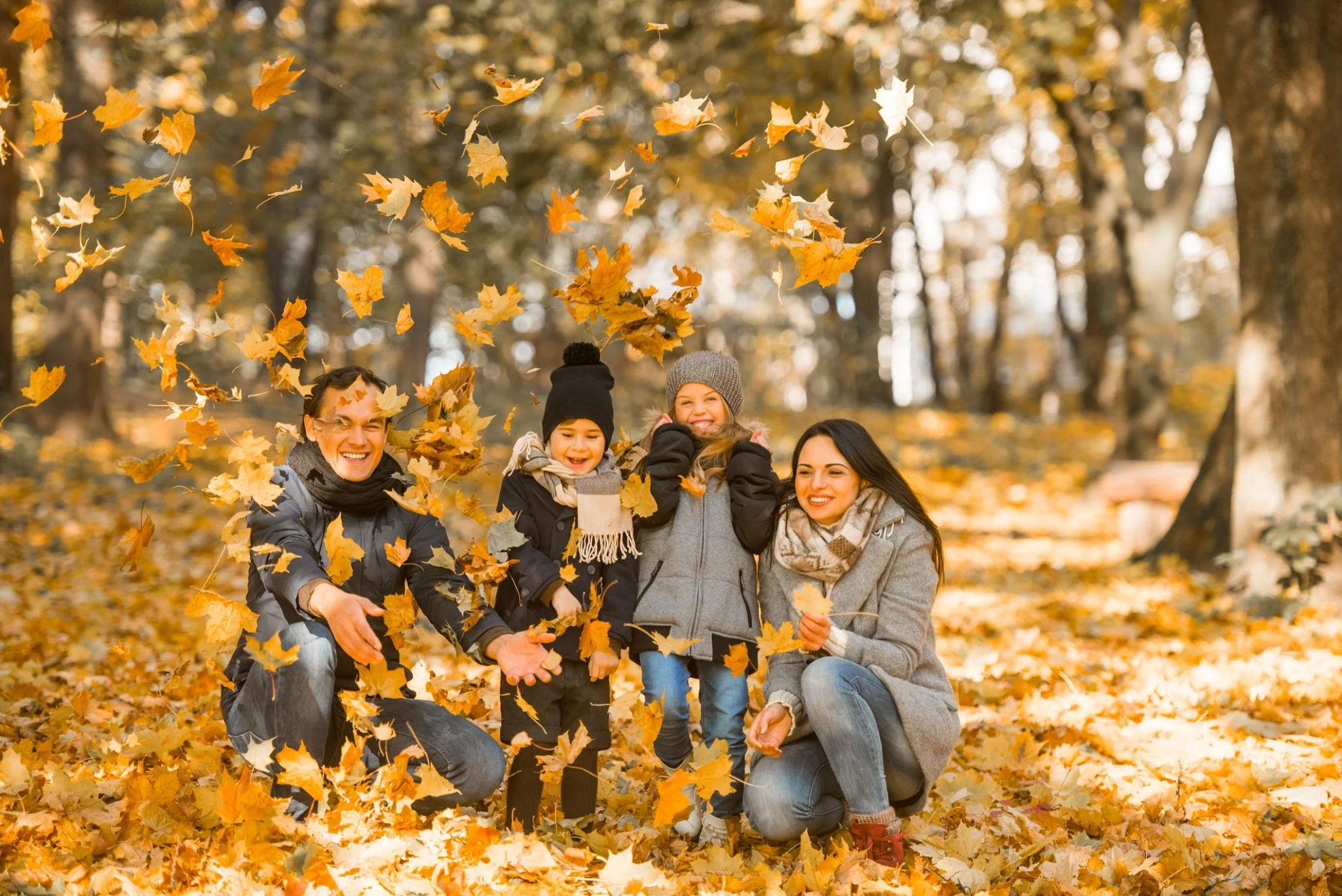 A family of five, including three children, is enjoying the fall weather among trees with yellow and orange leaves, some of which are being tossed in the air.