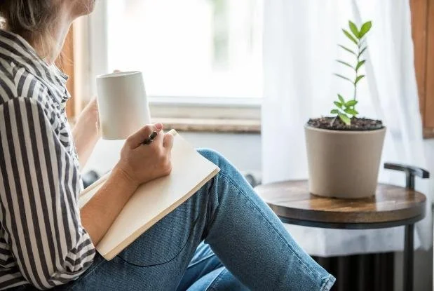 Person sitting on a windowsill holding a mug and writing in a notebook, with a potted plant on a table nearby.