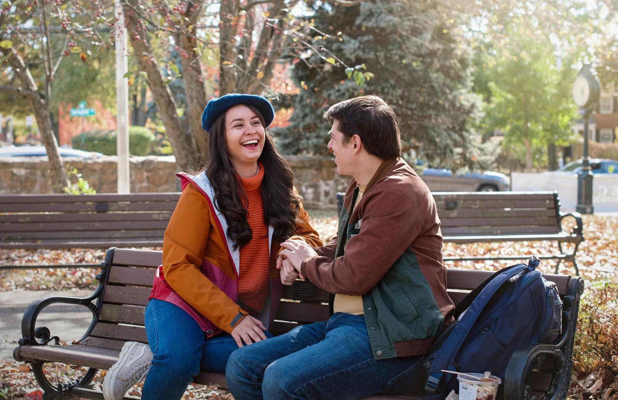 A young woman and a young man are sitting on a park bench, holding hands and smiling at each other. The woman wears a blue beret and an orange jacket, while the man wears a brown jacket. They are surrounded by trees with fall foliage, and there are other park benches and a blurred background of parked cars and buildings.