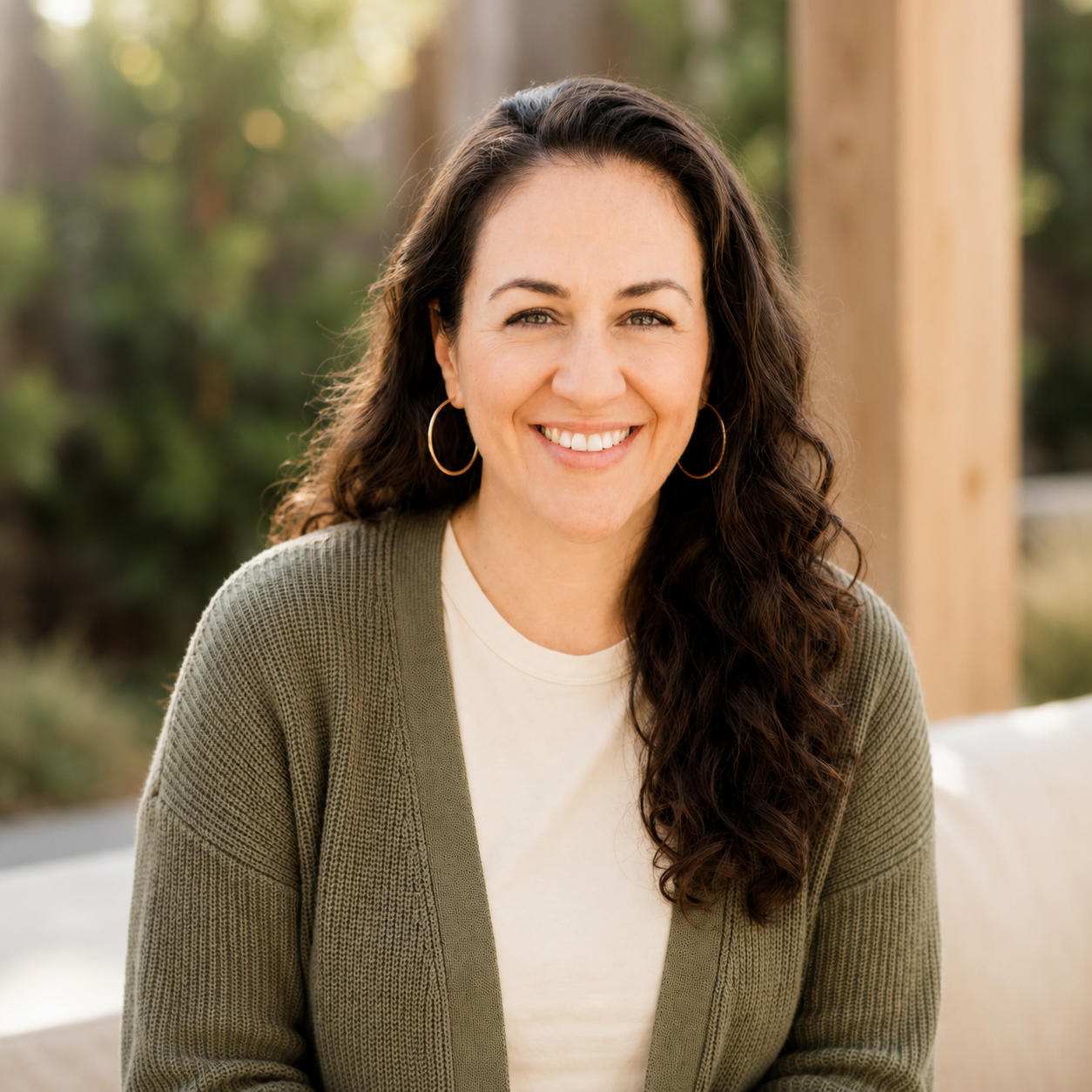 A woman with long, curly dark hair smiling outdoors, wearing hoop earrings, a white shirt, and a green cardigan.