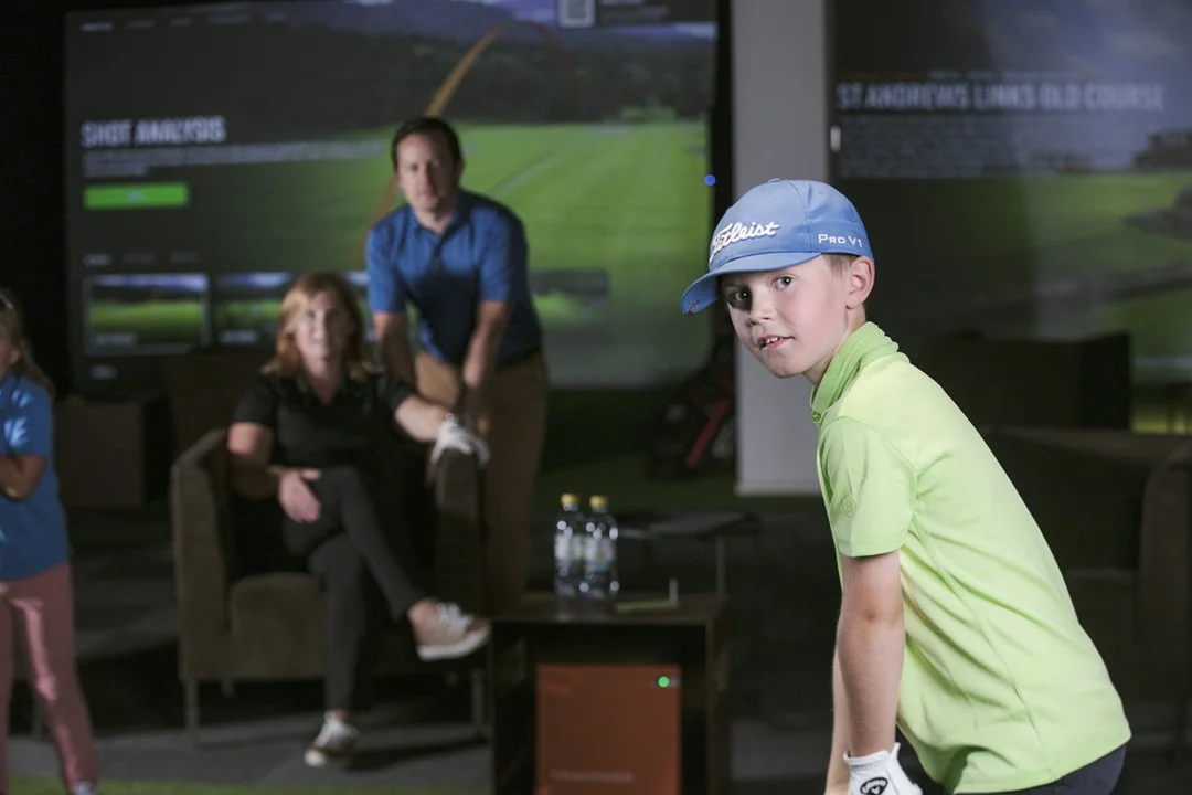 Young boy wearing a blue cap and a light green polo shirt at an indoor golf simulation game with others in the background.