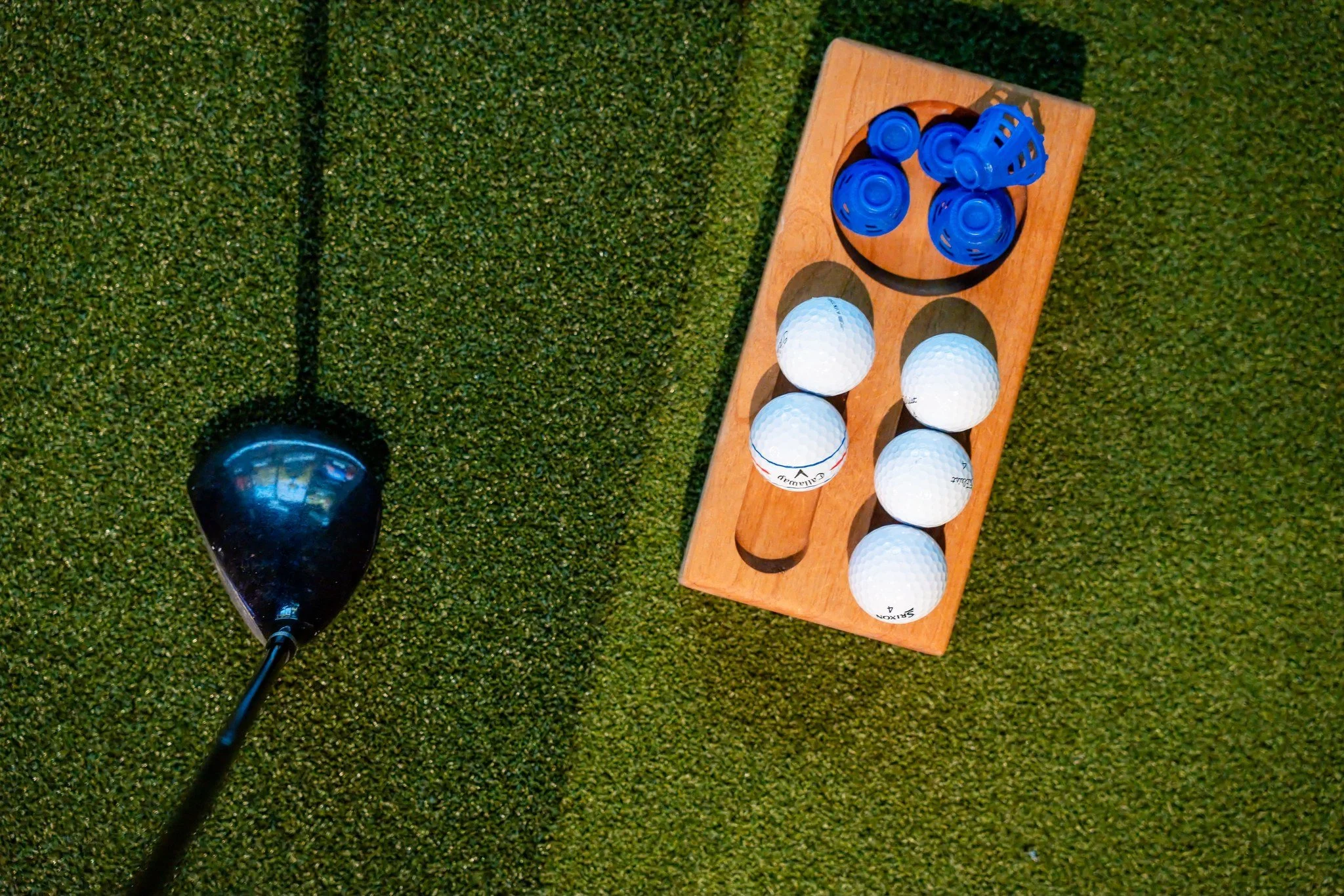 Close-up of a golf club and a wooden rack holding six golf balls on artificial grass, with a golf tee nearby.